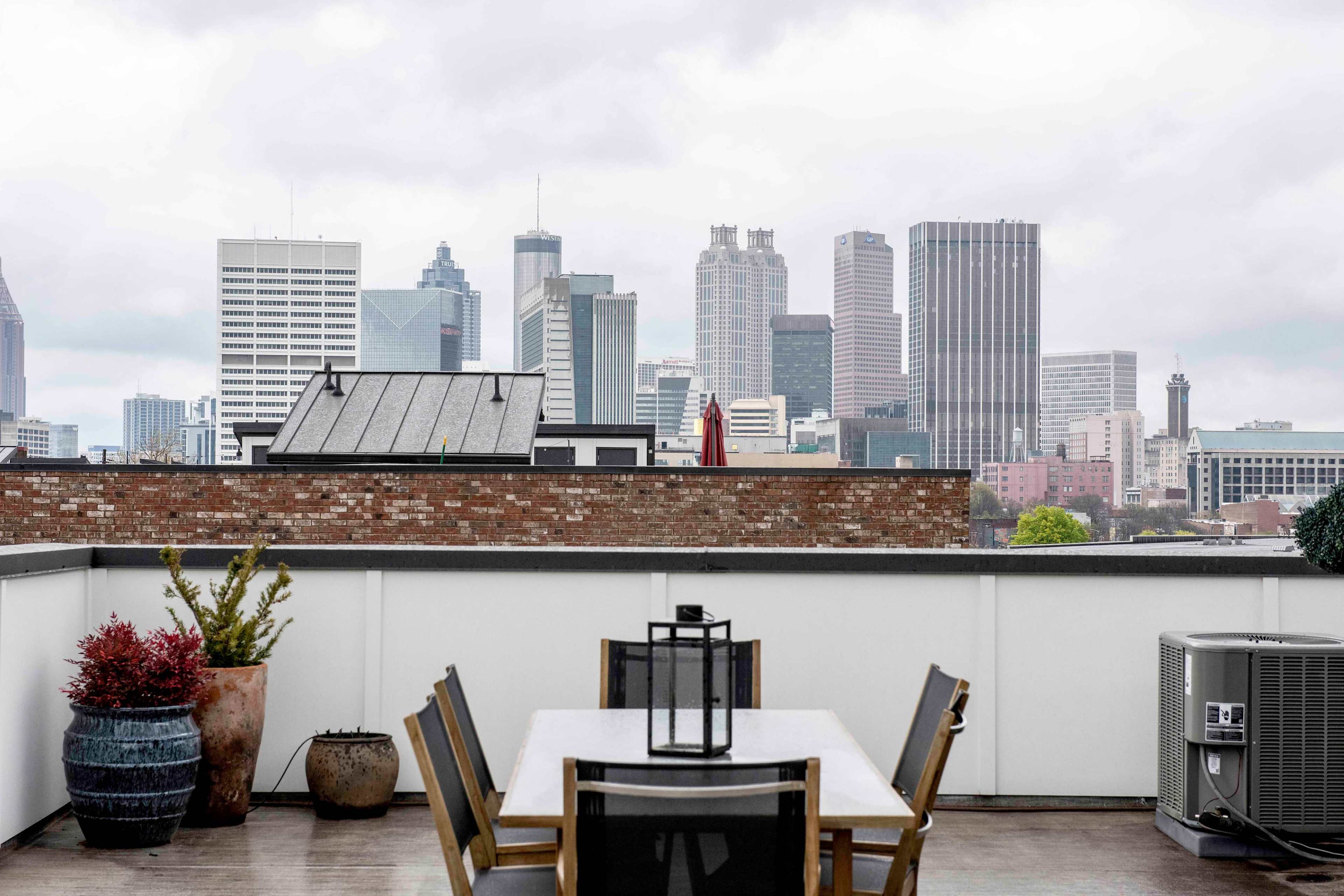 A rooftop terrace features a dining table and chairs, overlooking a city skyline with tall buildings under a cloudy sky.
