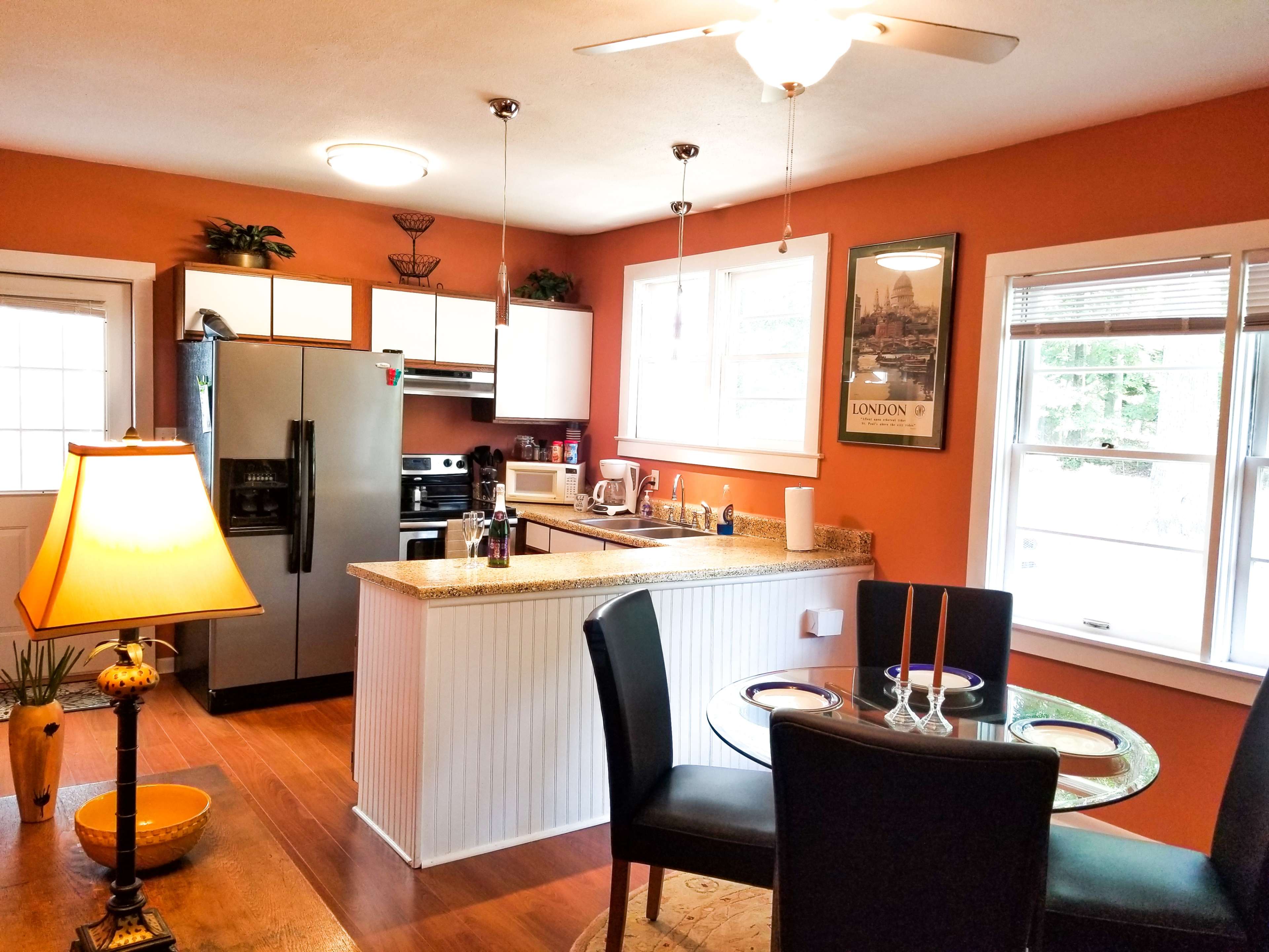 A kitchen with orange walls, modern appliances, a dining table for two, and natural light from adjacent windows.