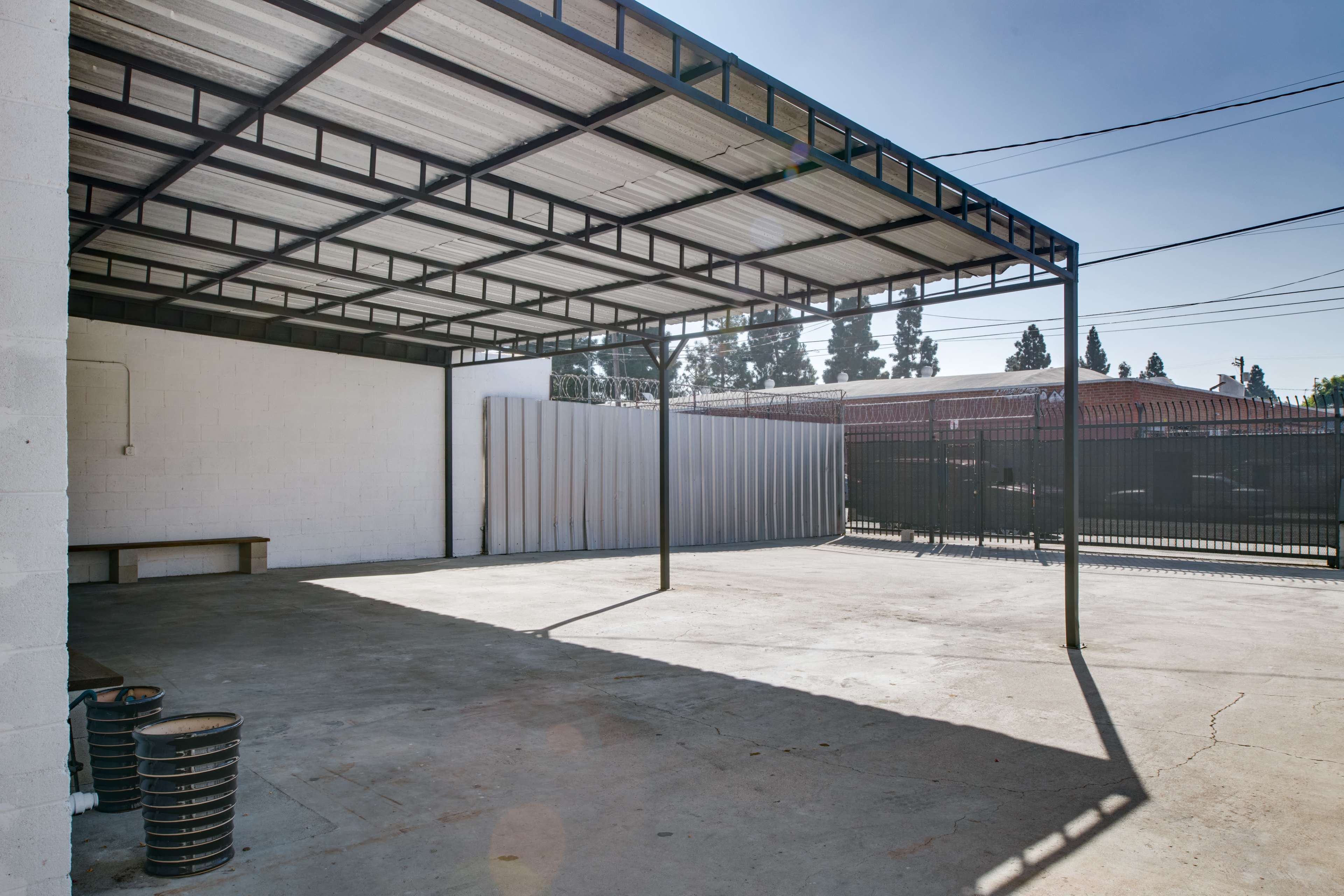 An empty outdoor area beneath a metal canopy, with concrete flooring and adjacent walls.