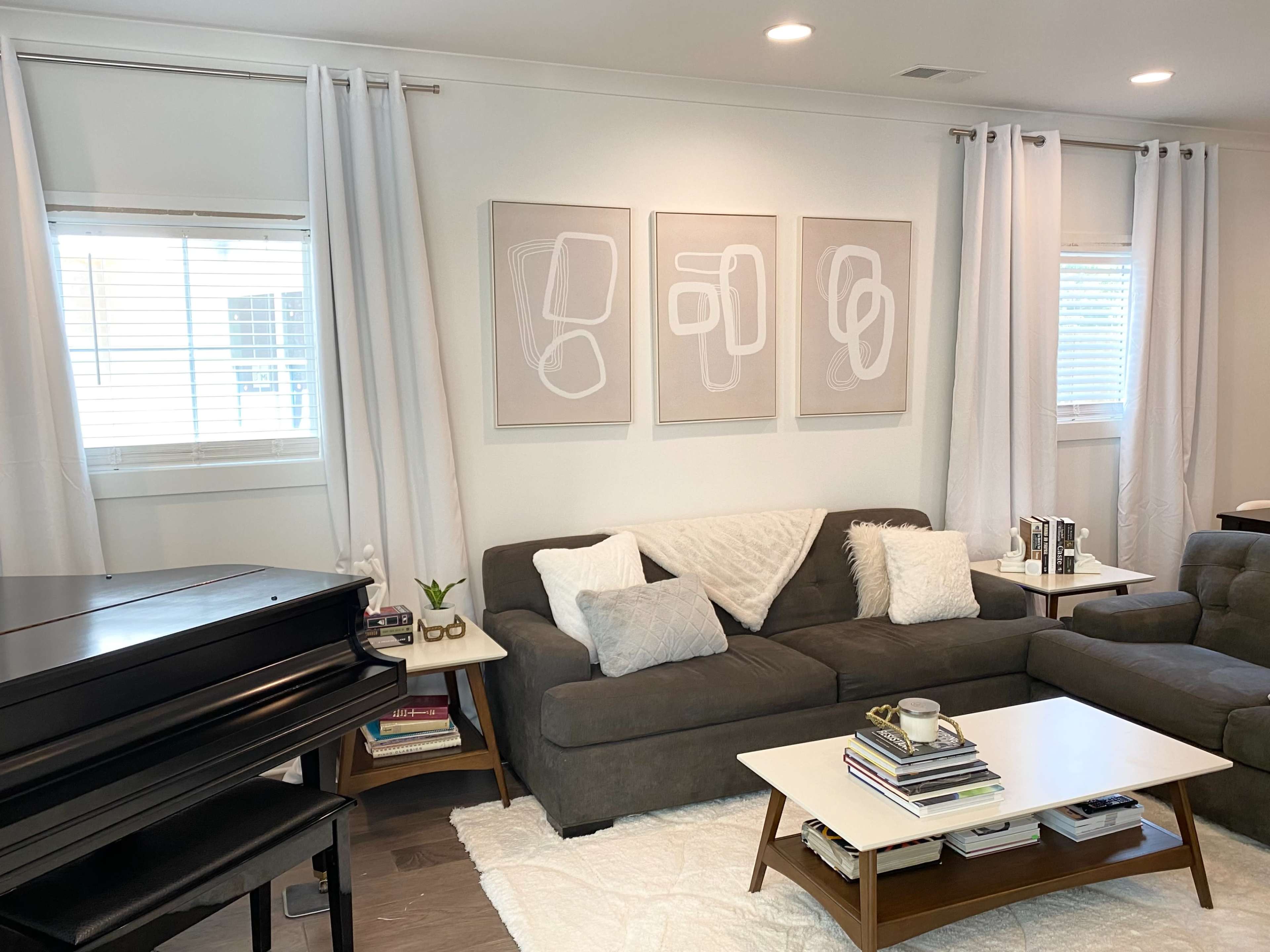 The image shows a modern living room featuring a dark sectional sofa, a coffee table with books and a plant, a grand piano in the corner, and three framed abstract artworks on the wall.