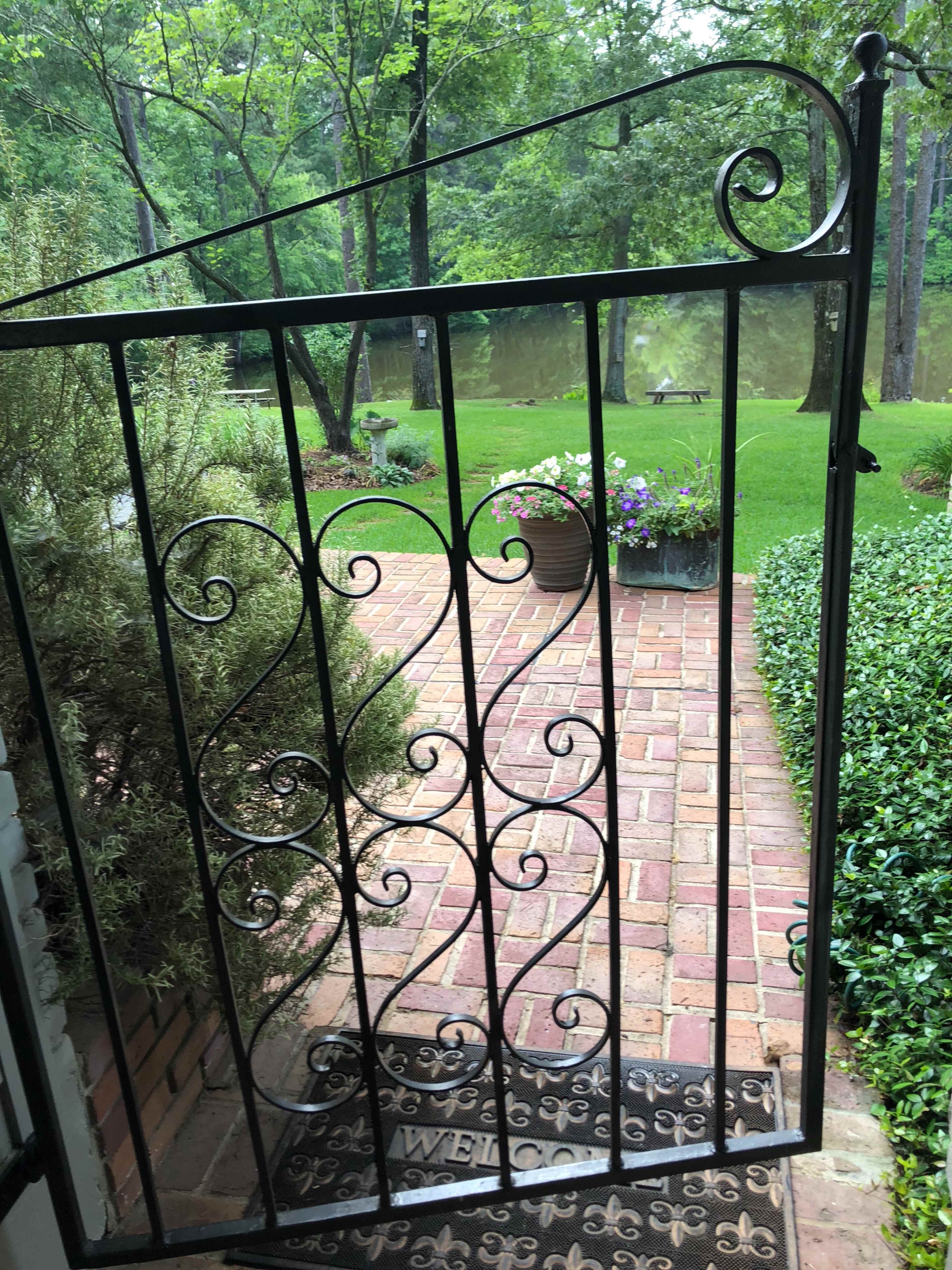 An ornate metal gate opens onto a brick pathway lined with greenery and flower pots, leading to a tranquil area near a body of water.
