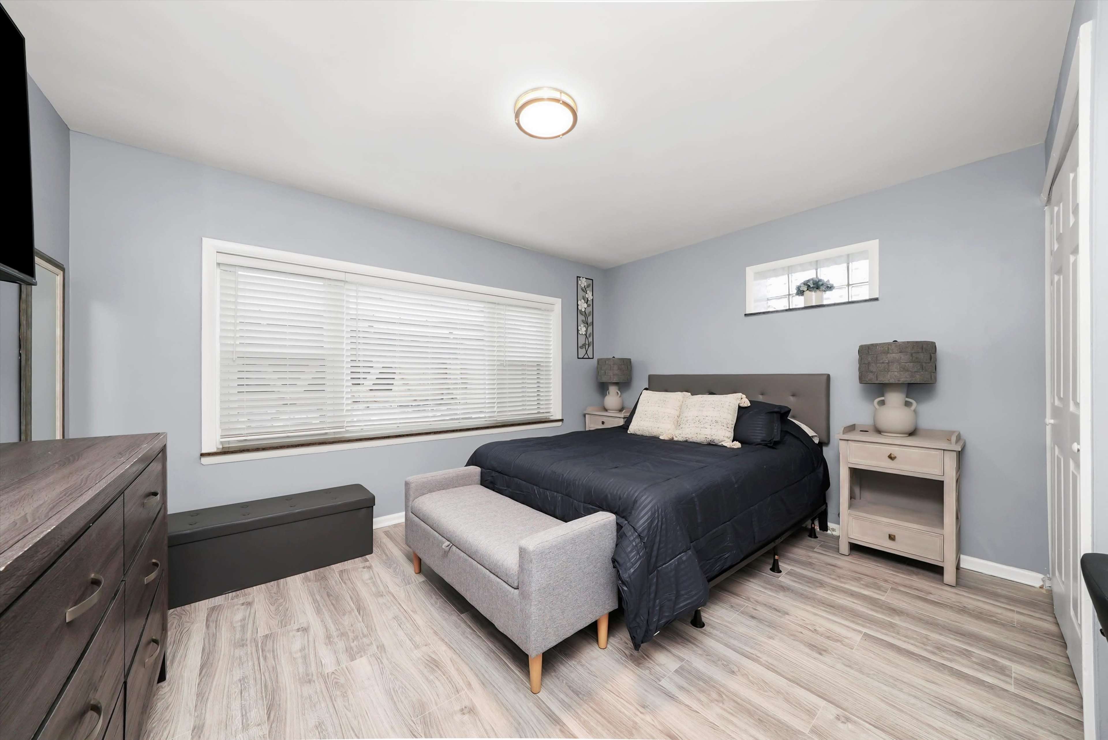 The image shows a neatly arranged bedroom featuring a bed, two bedside tables with lamps, a dresser, and a large window with blinds.