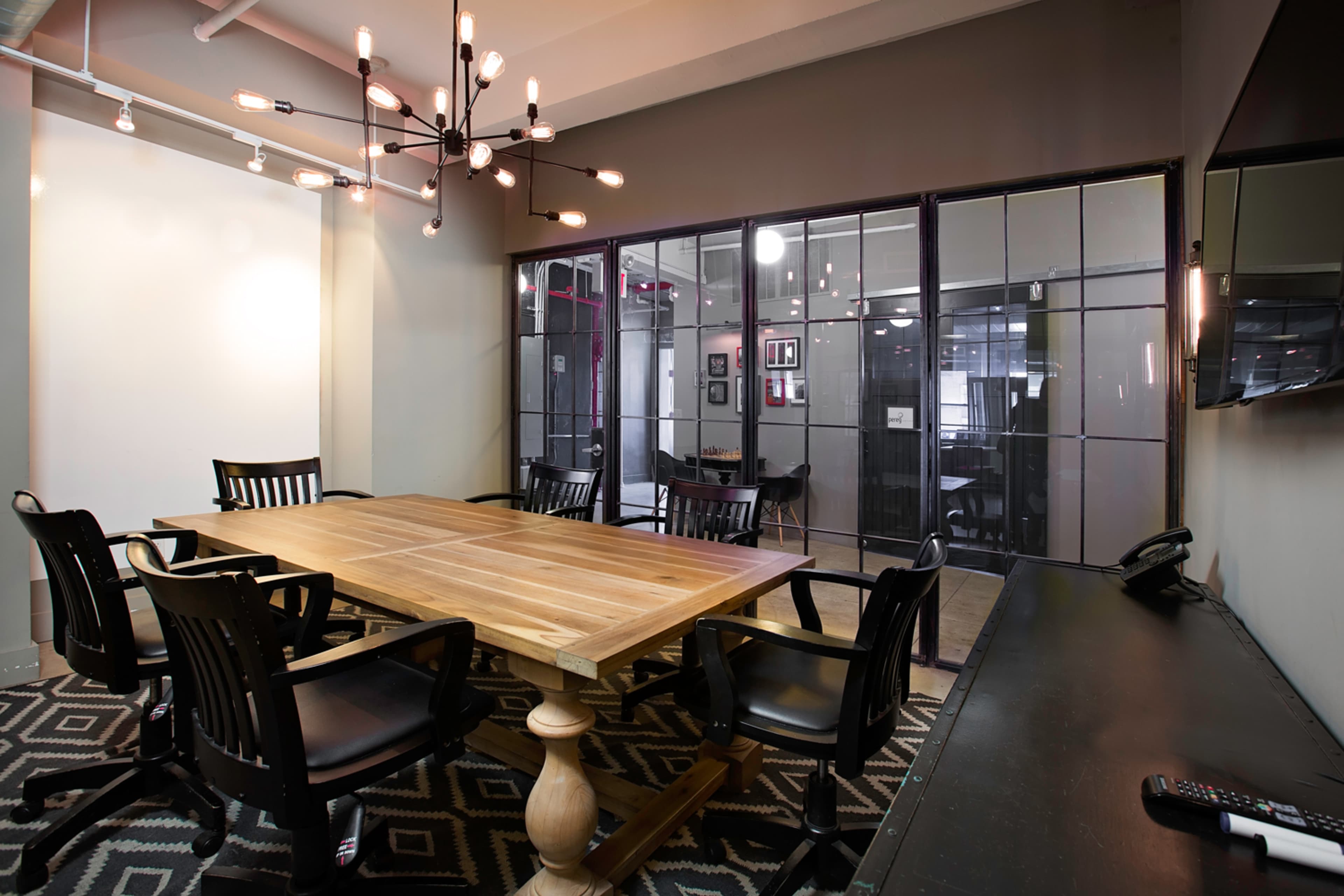 The image shows a modern conference room with a wooden table surrounded by black chairs, featuring a light fixture and glass walls separating it from adjacent spaces.