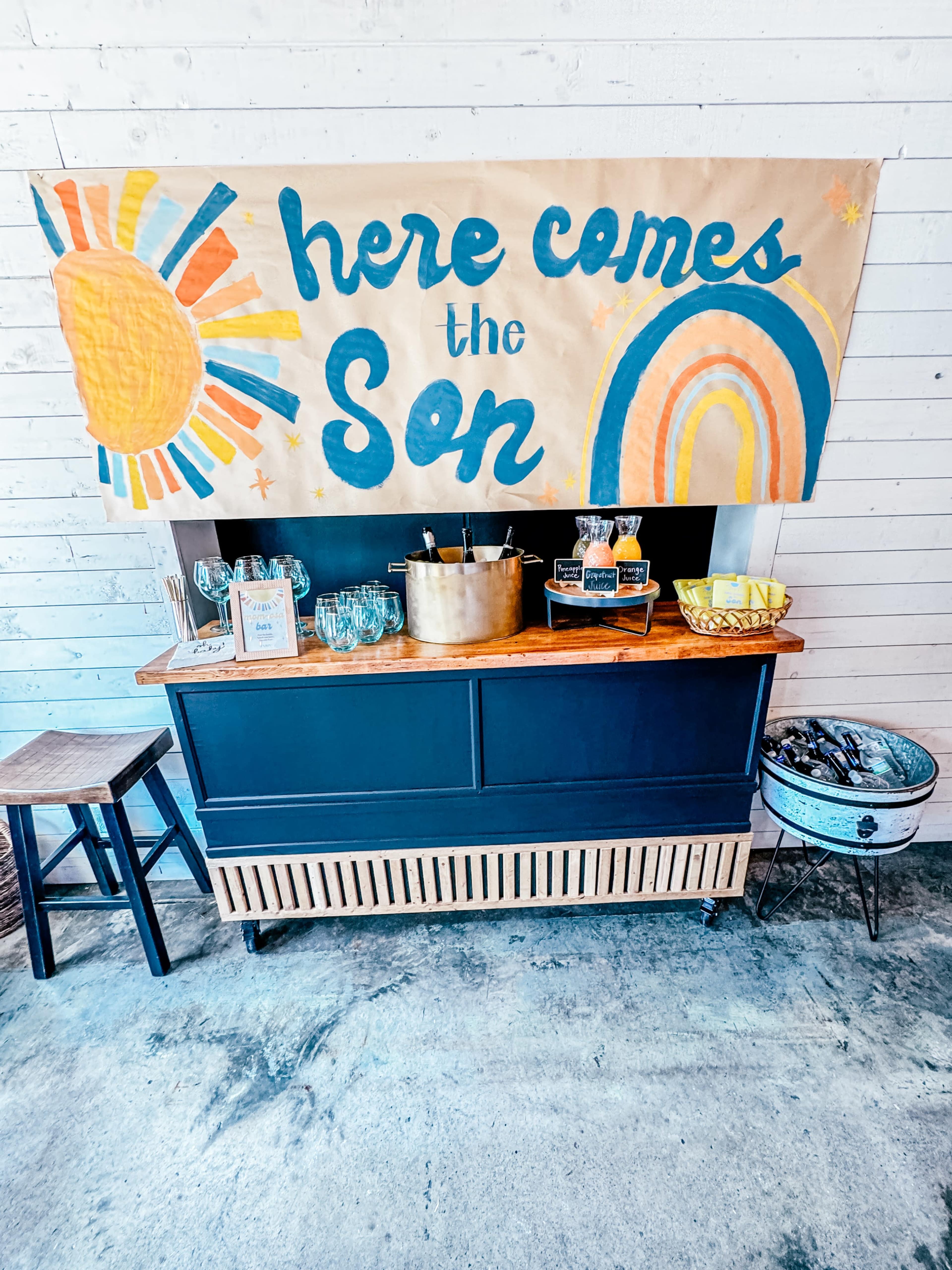 A table is decorated with a colorful banner reading "here comes the Son," with a sun and rainbow illustration, alongside a gold container and glassware.