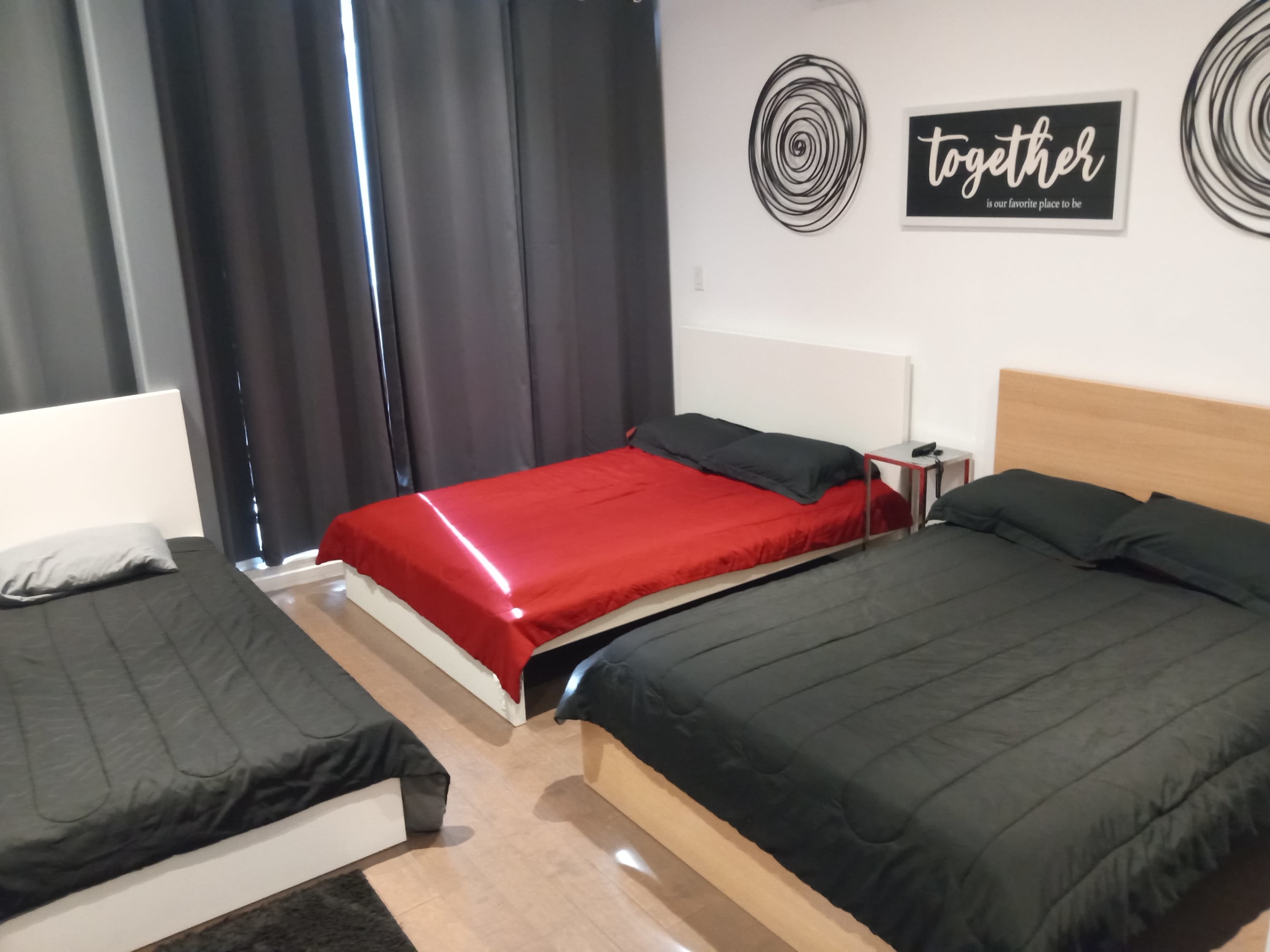 The image shows a room with three beds: one red and two black, along with grey curtains and a wall decoration that reads "together."