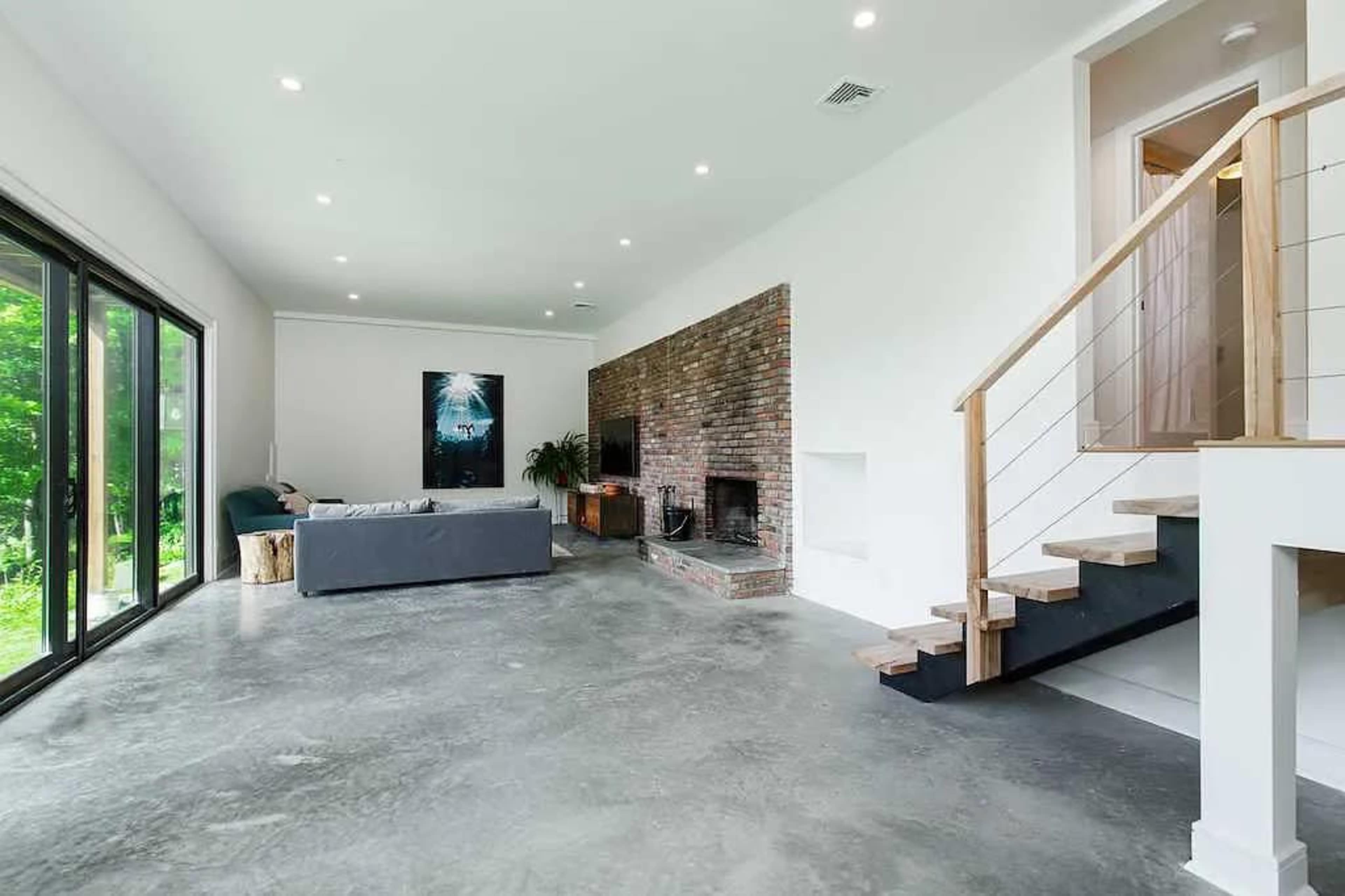 A spacious living room with a gray sofa, a brick fireplace, and large windows overlooking greenery.