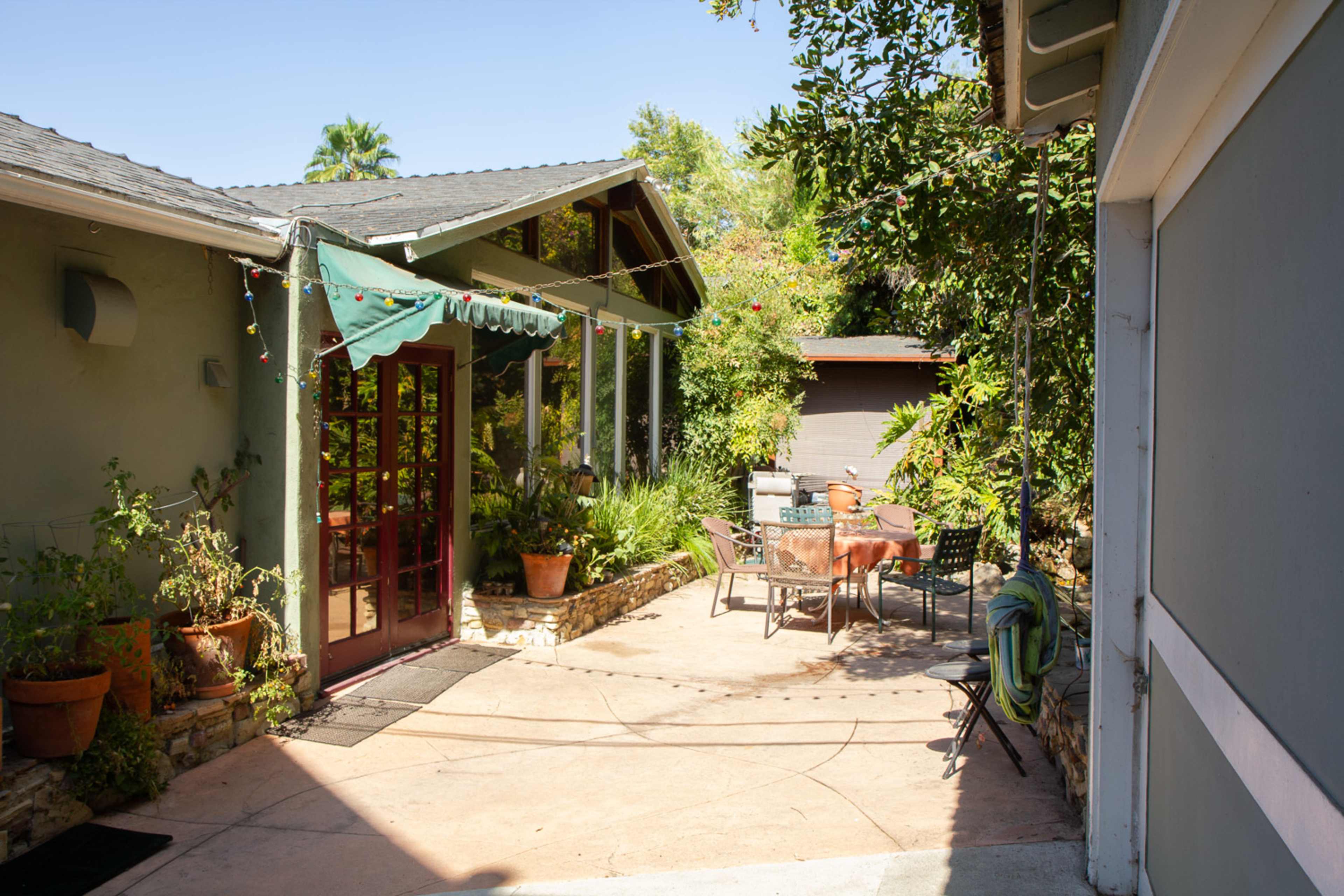 A sunny patio area with a table and chairs surrounded by plants and a house with large windows.