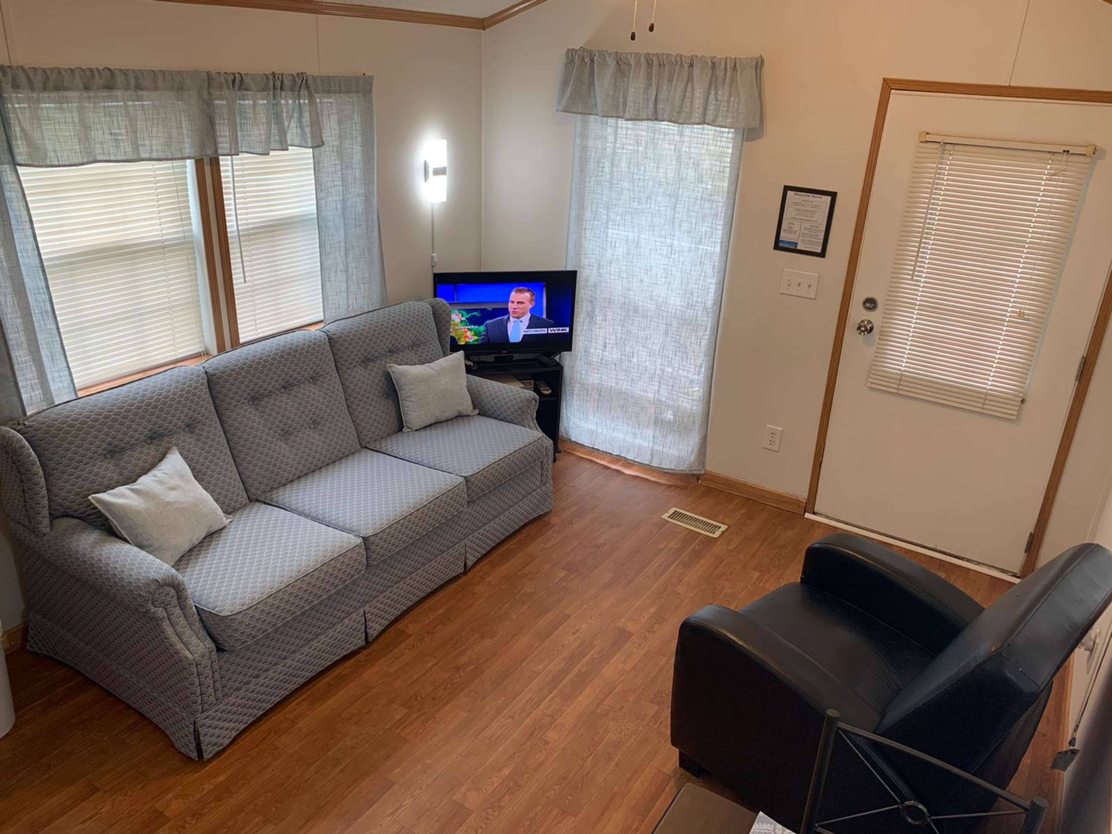 A living room features a gray couch with white pillows, a television in front showing a news program, and a black armchair near a door with a blind.