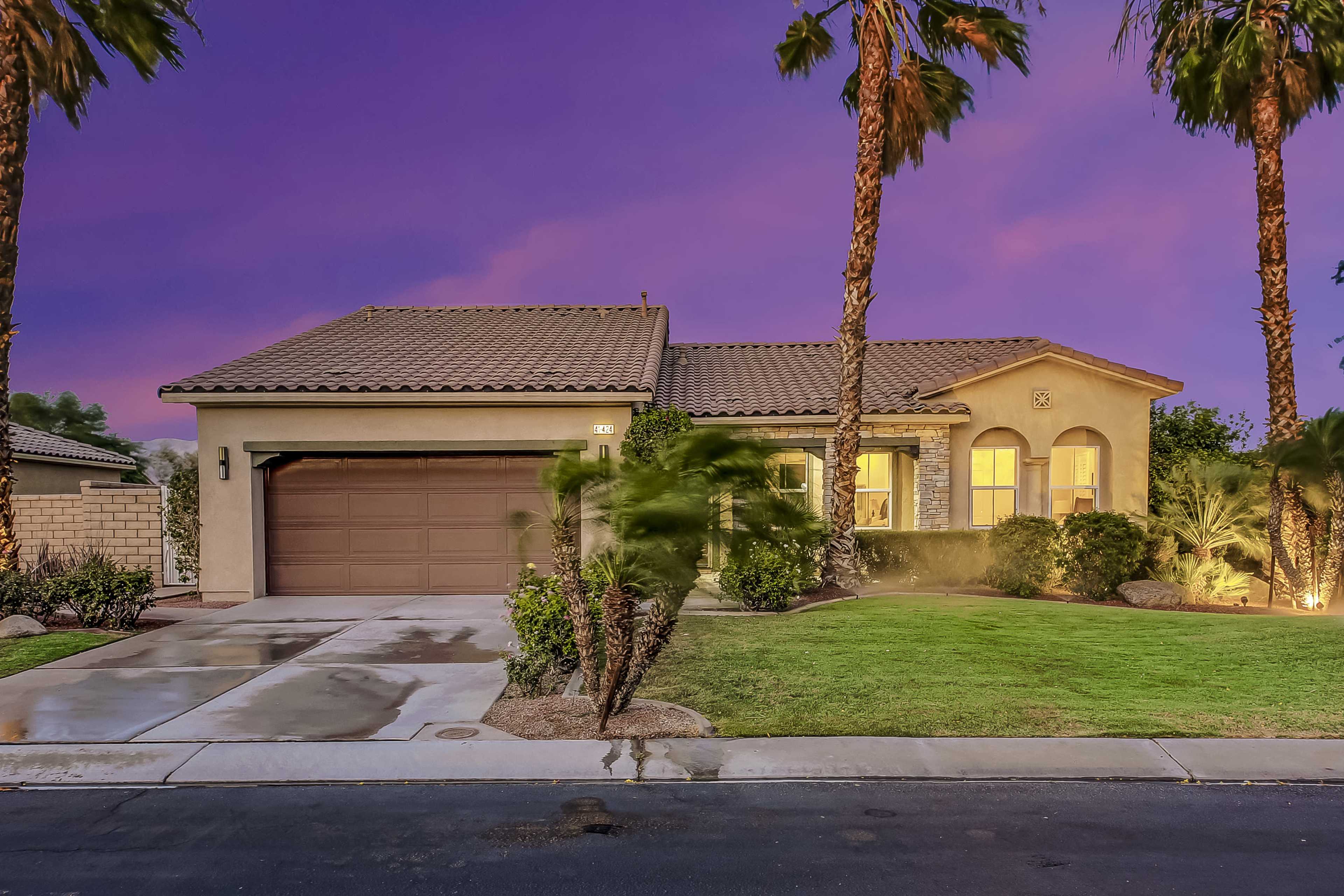 A single-story house with a tiled roof and a two-car garage is surrounded by palm trees and greenery under a purple sky at dusk.