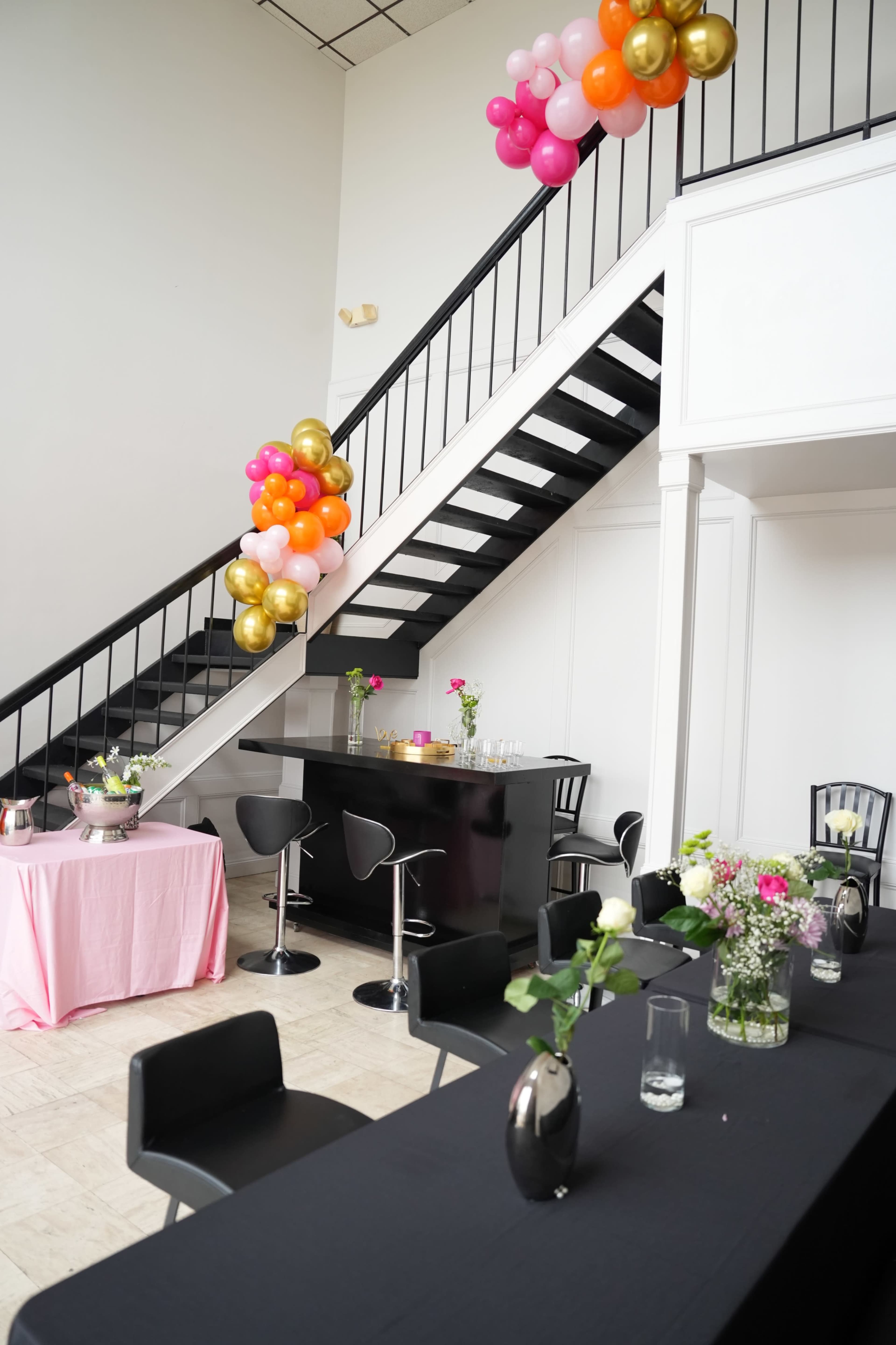 The image shows an indoor event space with a black bar, decorated tables, and colorful balloon arrangements along a staircase.