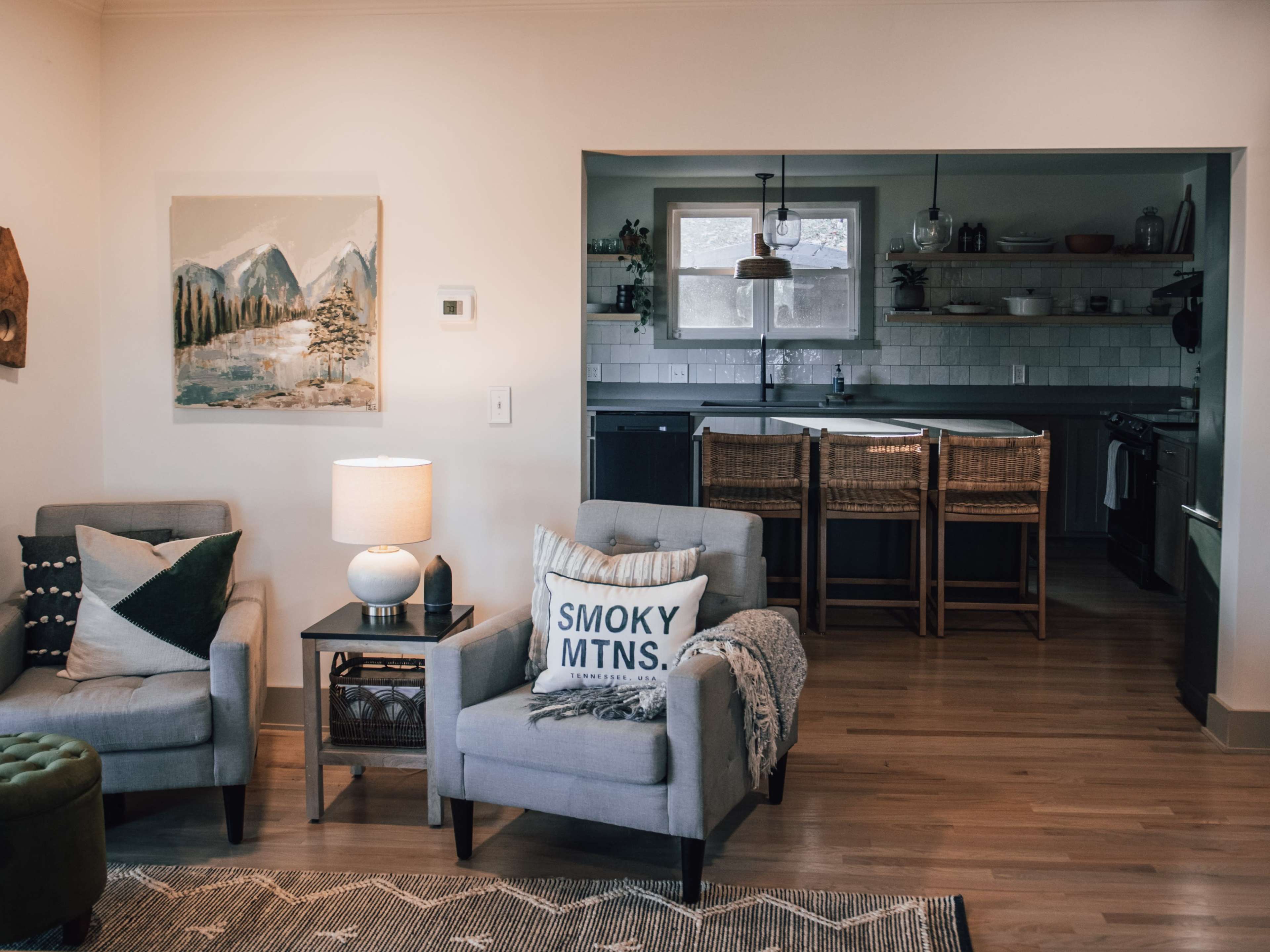 A cozy living room with two armchairs, a lamp, and a painting on the wall, leading to an open kitchen with wooden shelves and bar stools.