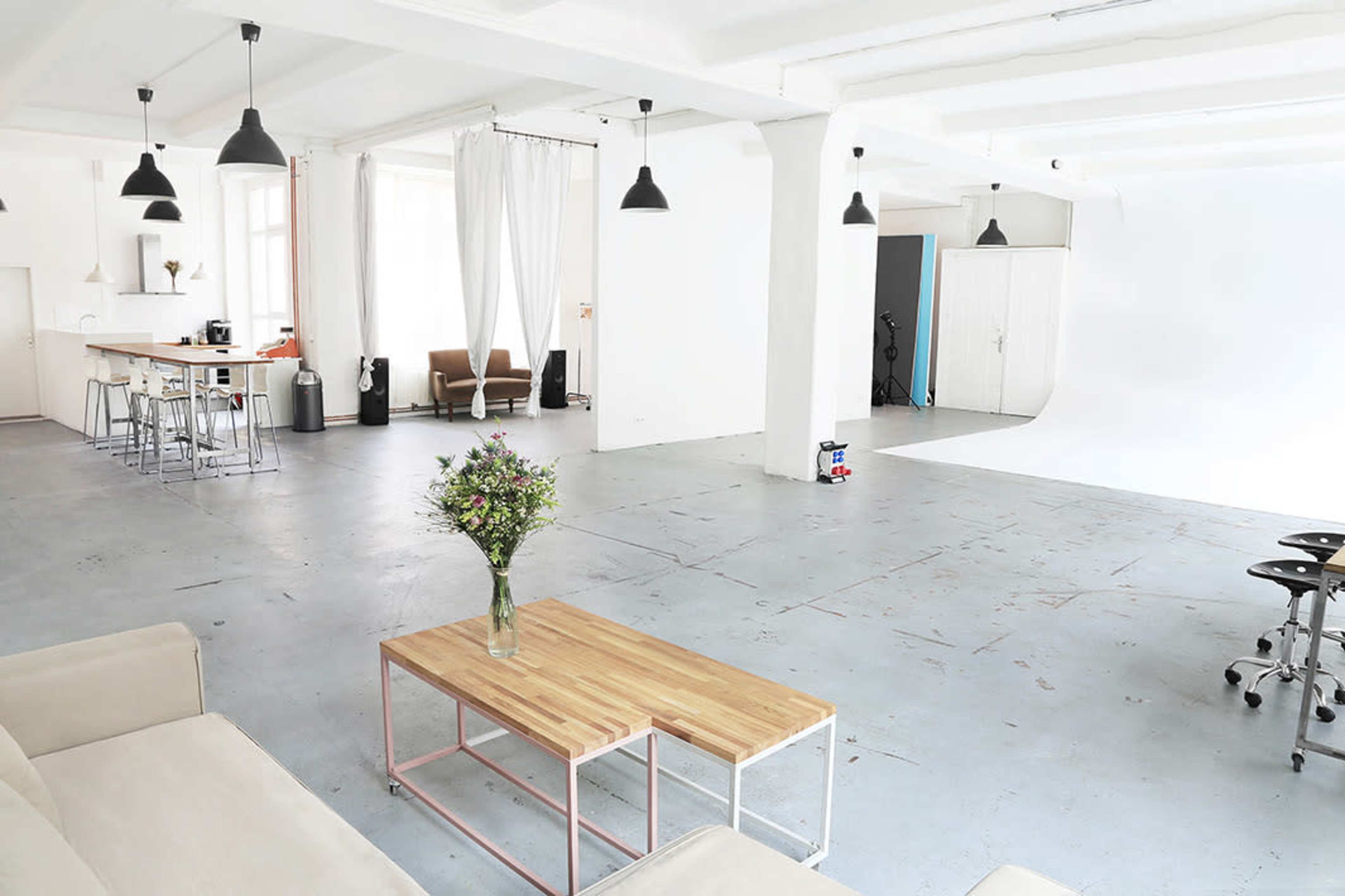 A spacious, well-lit studio with white walls, two wooden coffee tables, and a seating area, along with a photography backdrop on one side.