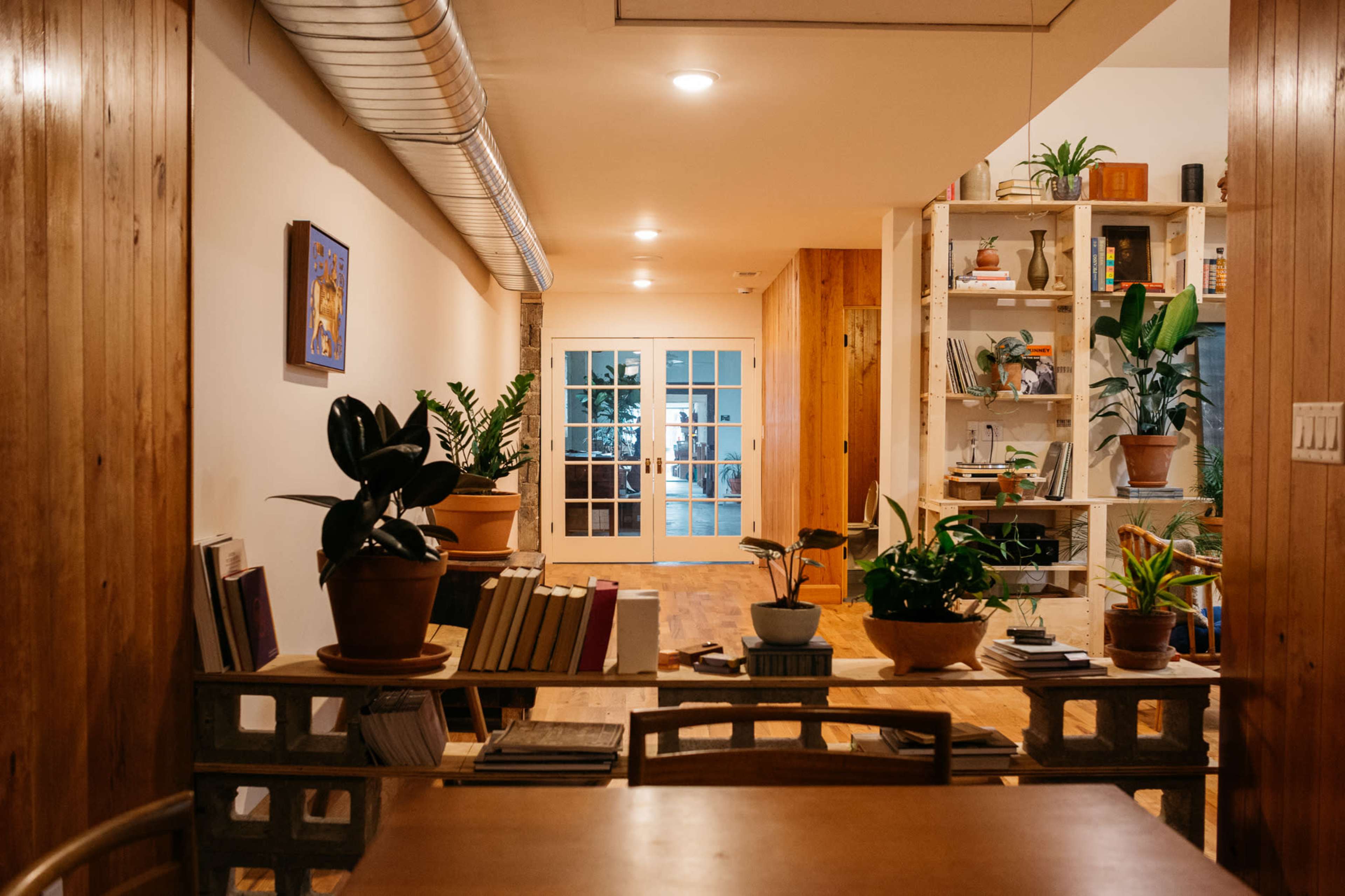 A warmly lit interior space with wooden accents, featuring a dining table in the foreground, plants on shelves, and a pair of double doors leading to another room.
