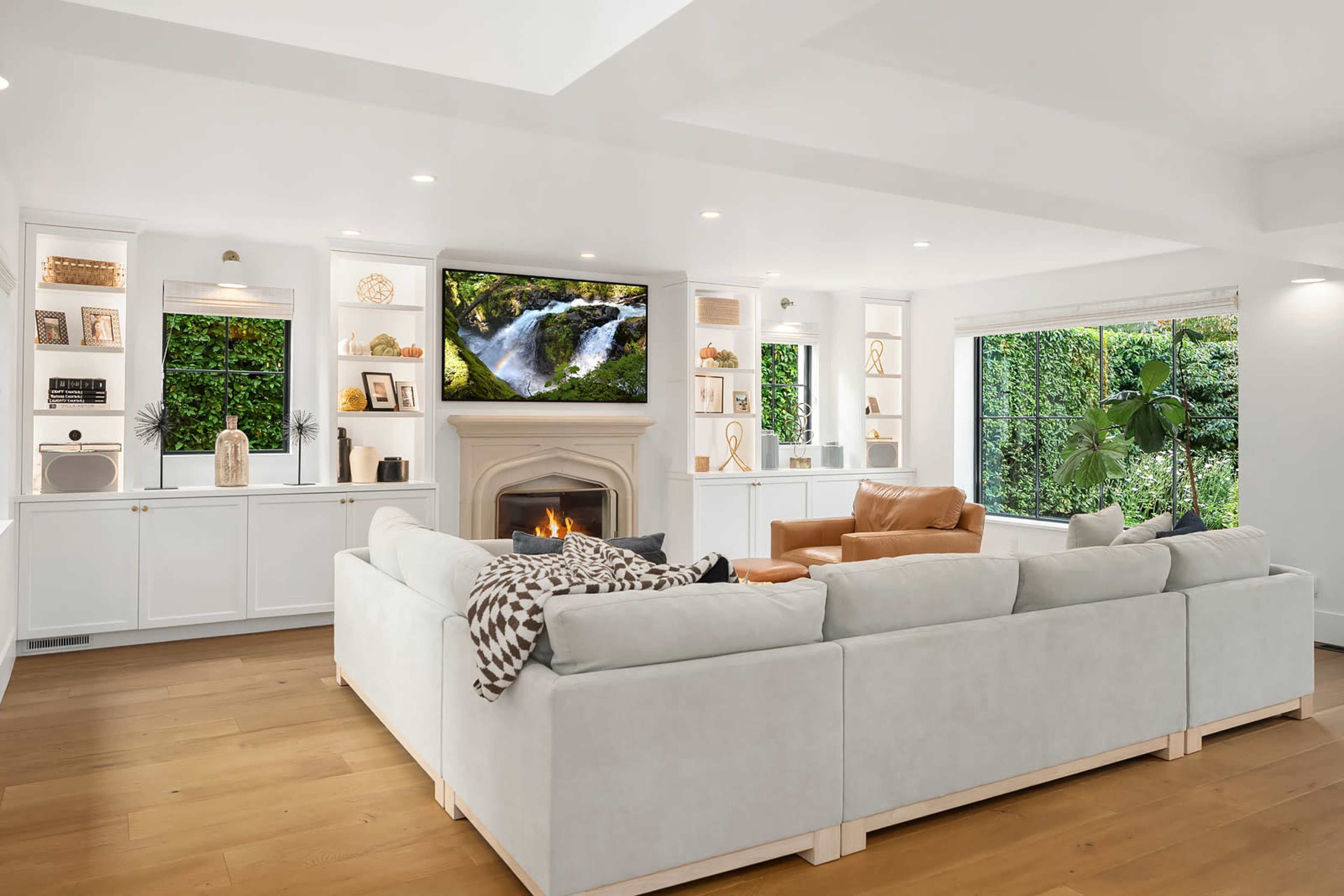 A modern living room featuring a white sectional sofa, a fireplace, built-in shelves, a large TV, and windows overlooking greenery.