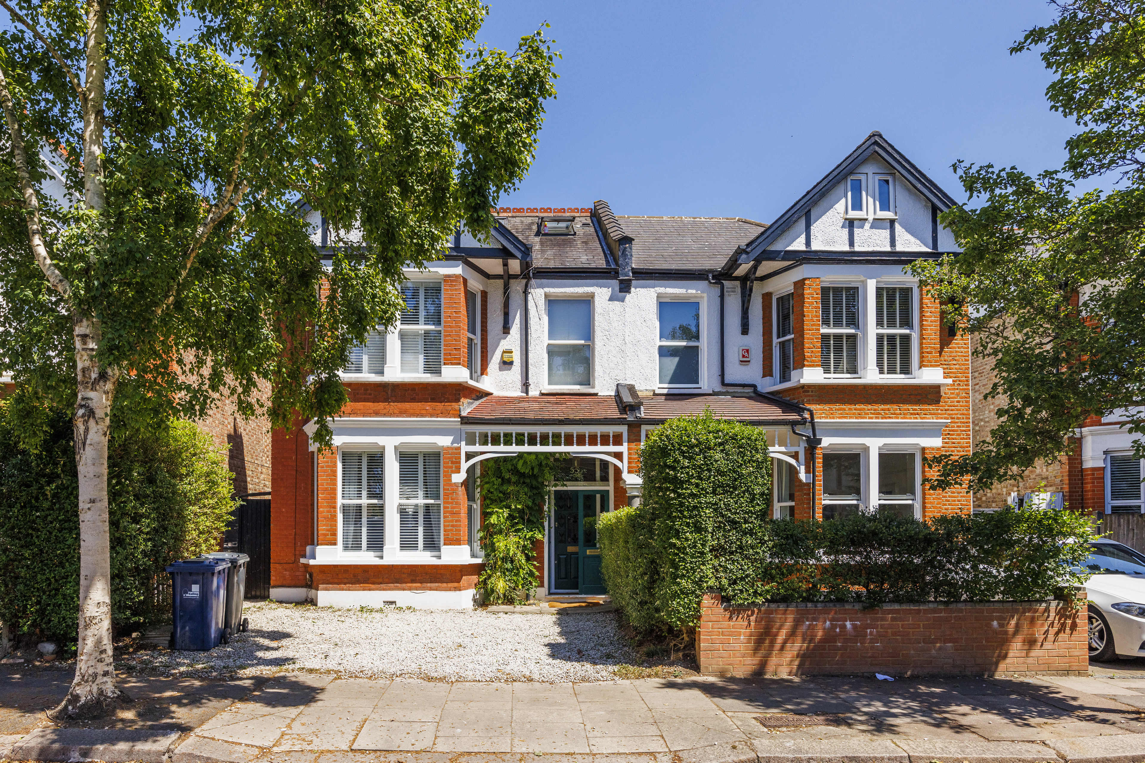 The image shows a two-story red-brick house with white trim, a gravel driveway, and lush greenery surrounding it.