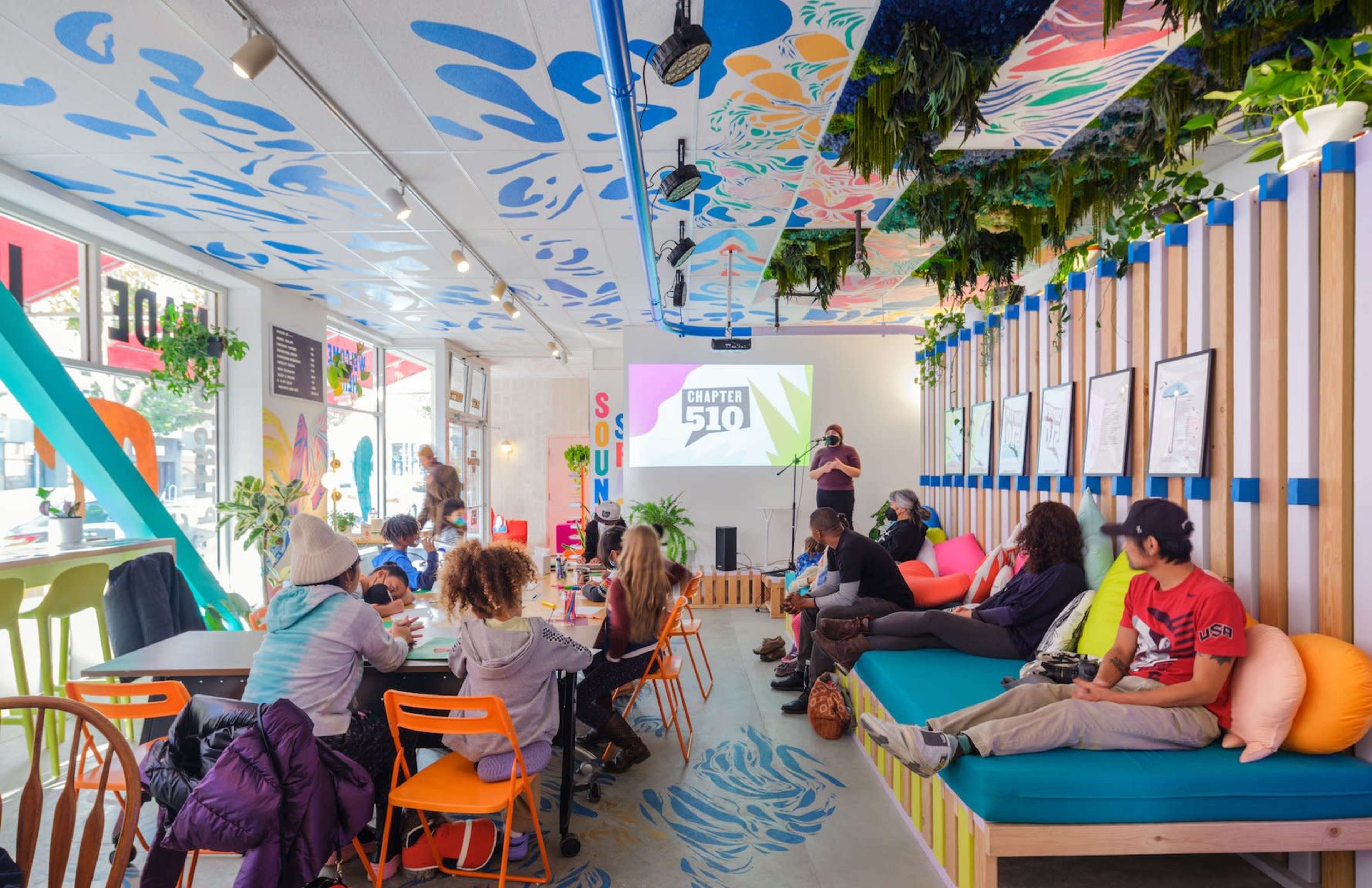 A lively café features colorful seating and a projector screen displaying "Chapter 510," while people engage in conversation and activities.