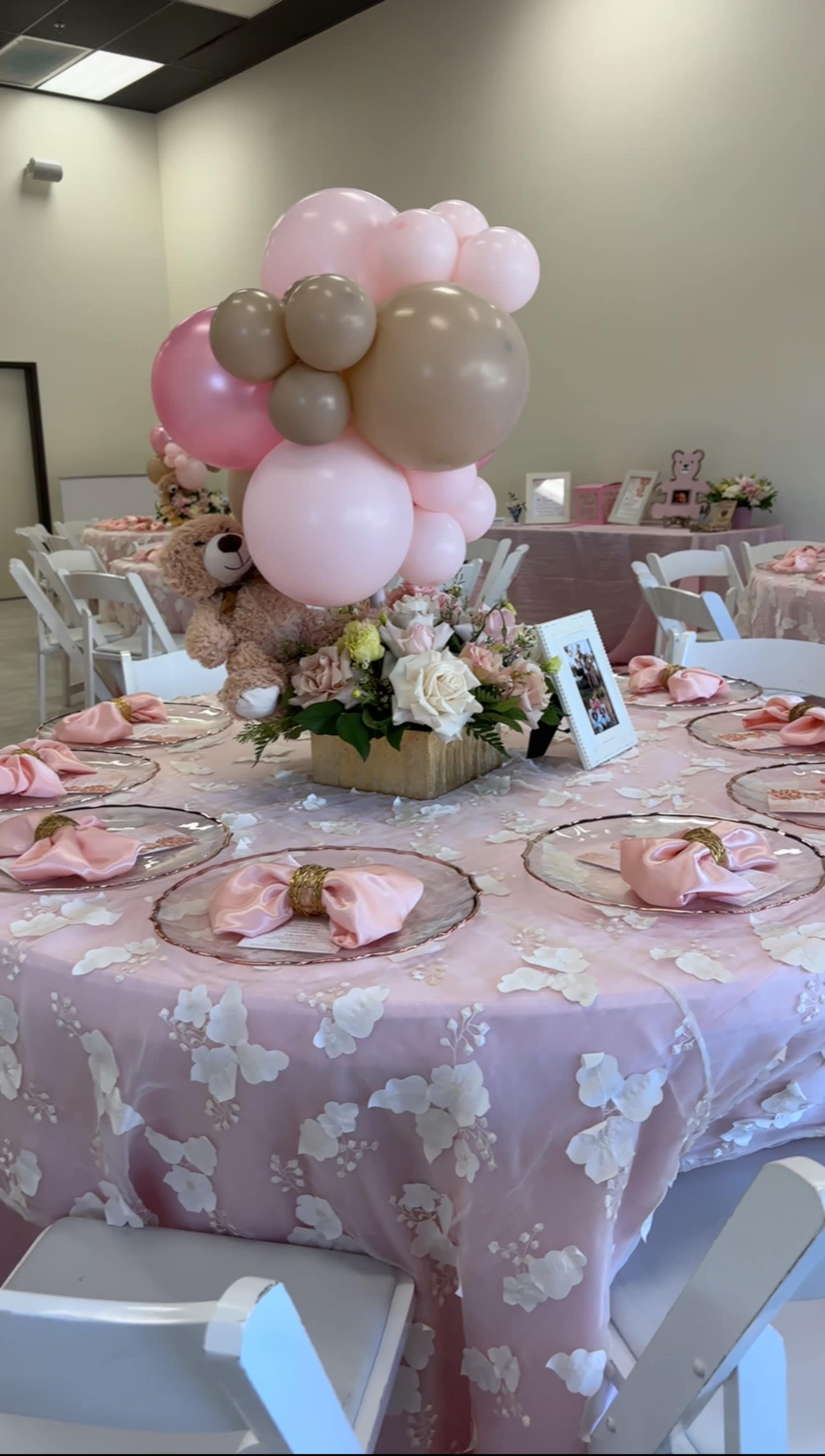 A decorated table features a pink floral tablecloth, glass plates, pink napkins, a floral centerpiece, and a cluster of balloons, with a teddy bear among the decorations.