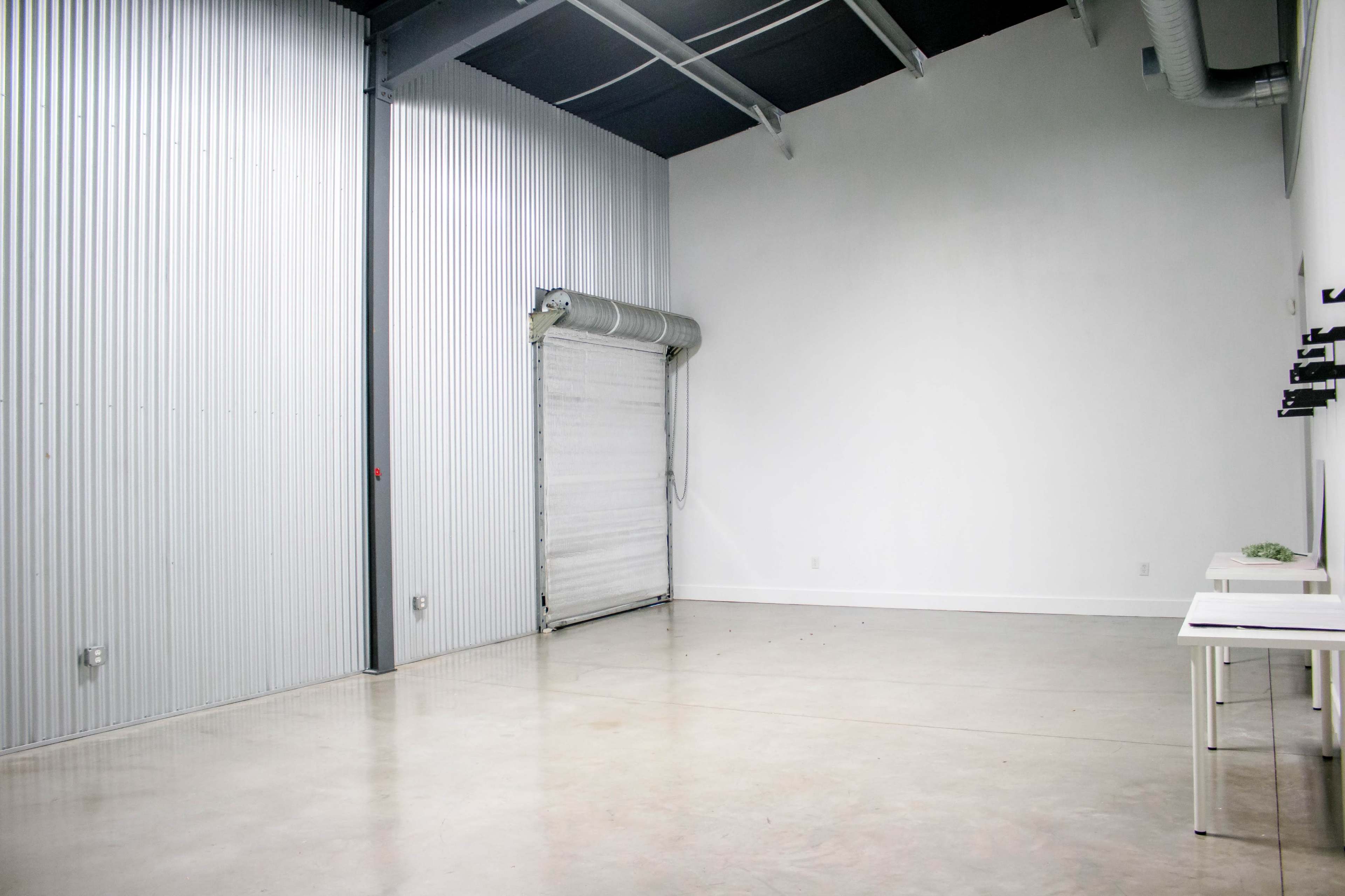 A spacious, industrial-style interior with metal walls, a large roll-up door, and empty tables against one wall.