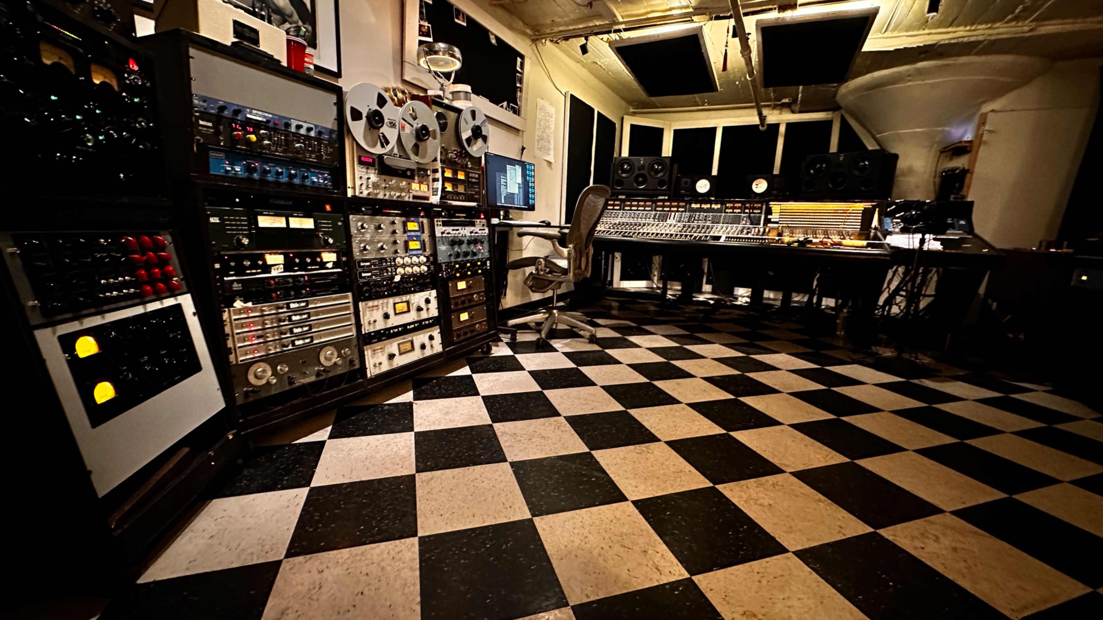 The image depicts a professional music studio with a checkered floor, various audio equipment racks, and a control desk equipped with monitors and mixing gear.