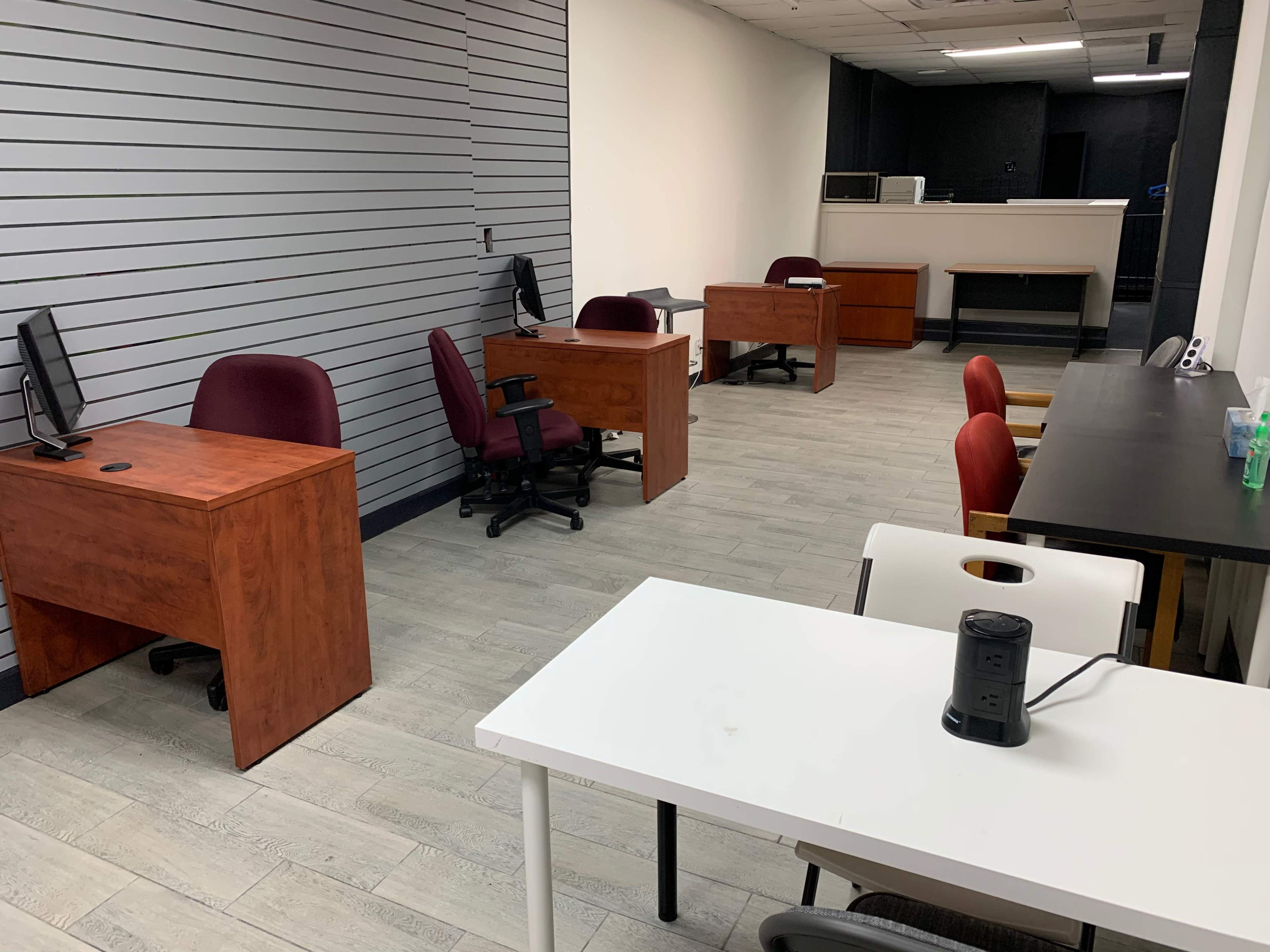 An office space with several wooden desks and chairs arranged in a mostly empty room with gray walls and a tiled floor.