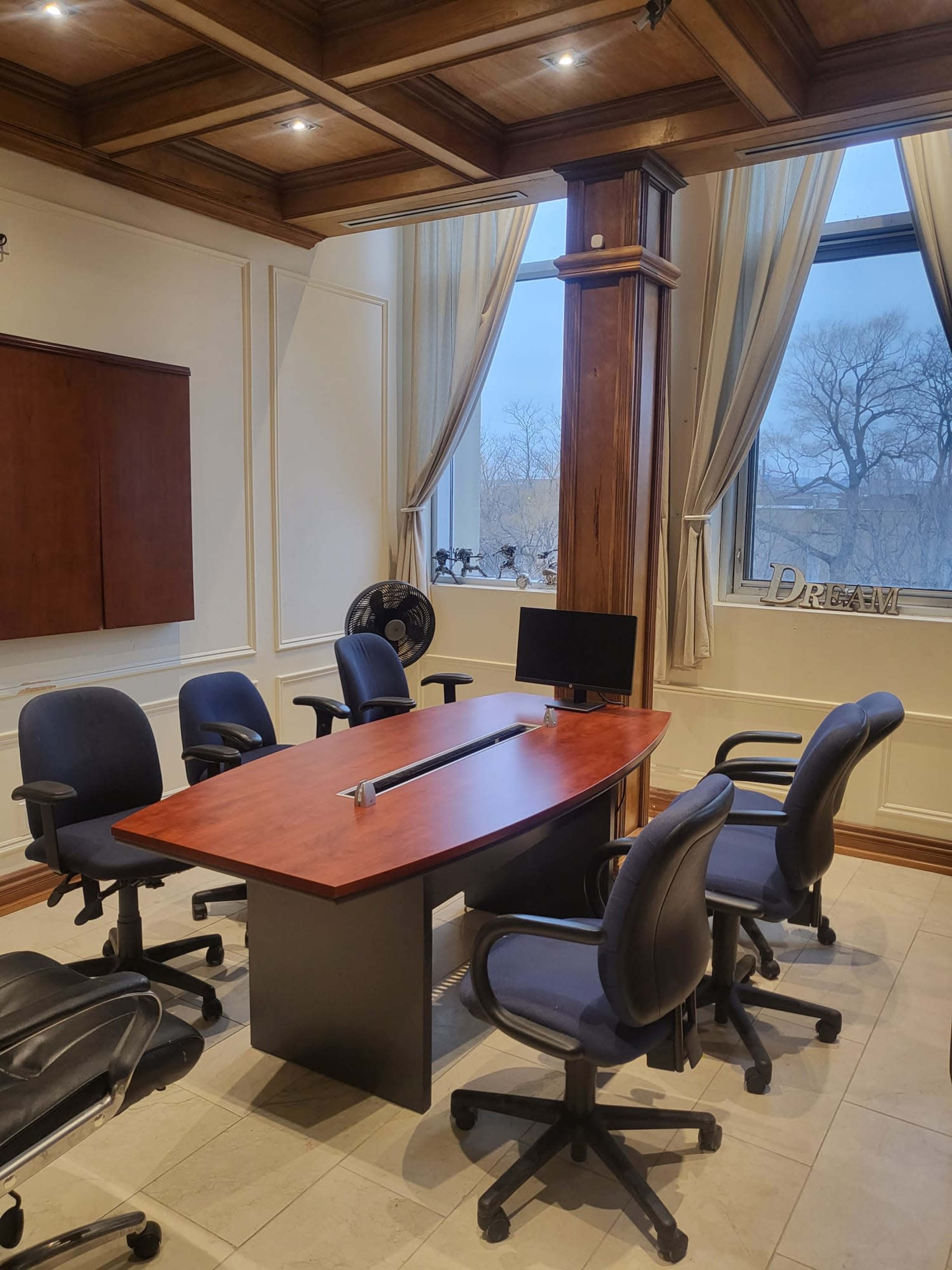 A conference room features a central wooden table surrounded by six office chairs, with a computer monitor and large windows overlooking a treed area.