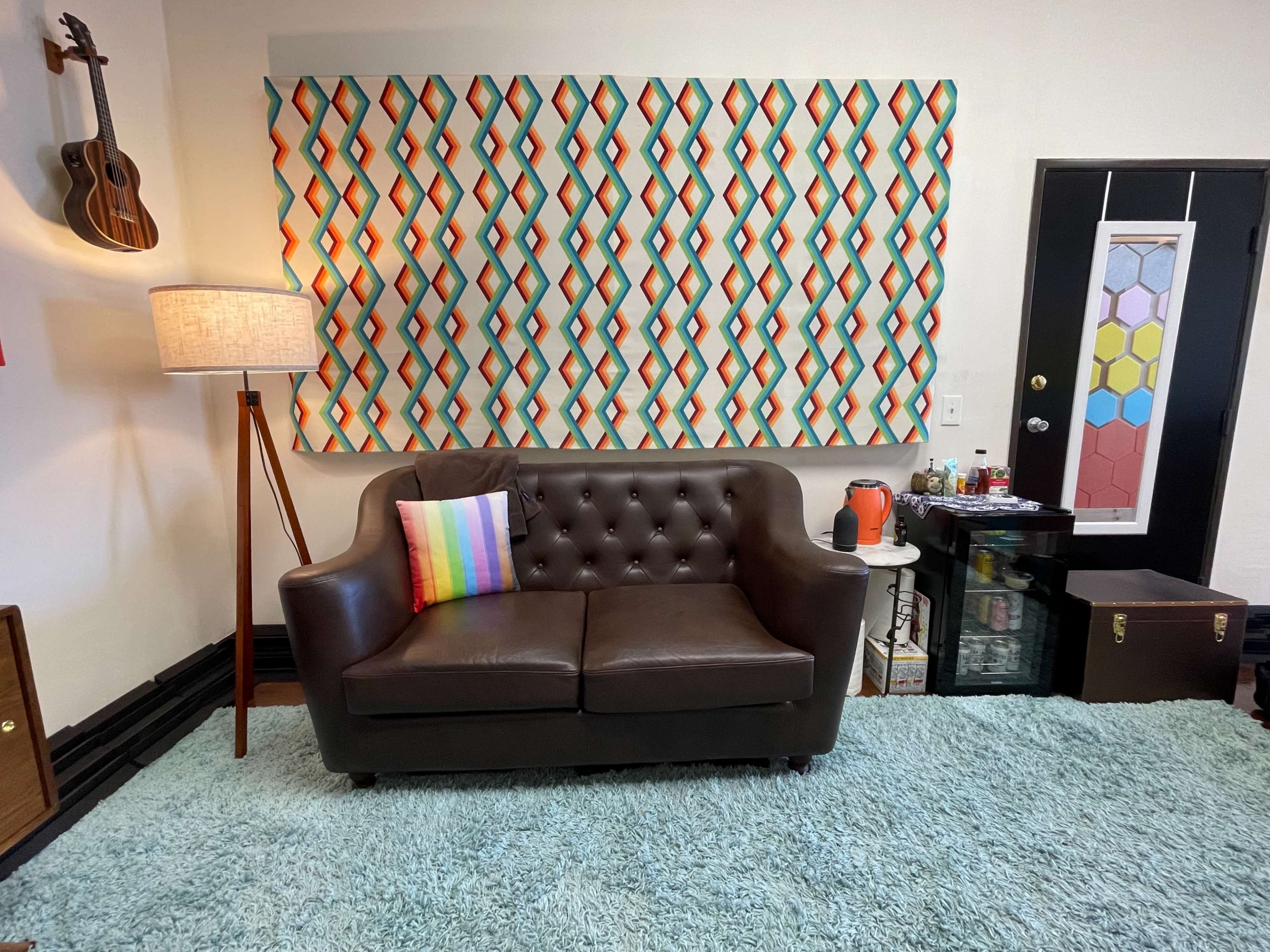 A living room with a brown leather couch, a colorful patterned wall hanging behind it, a standing lamp, and a teal rug on the floor.