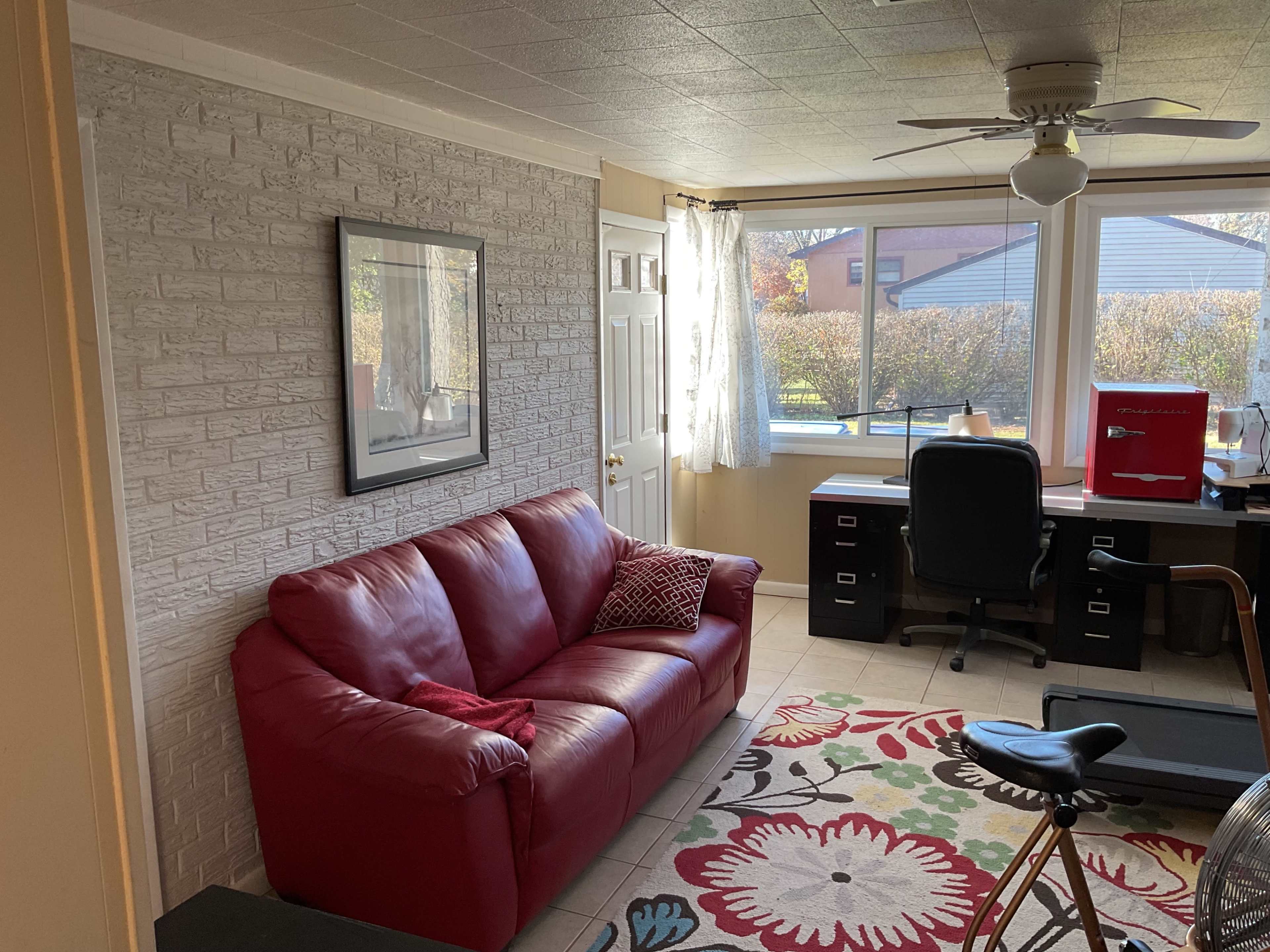 The image shows a cozy room with a red sofa, a desk with a chair, and a patterned rug, illuminated by natural light from large windows.
