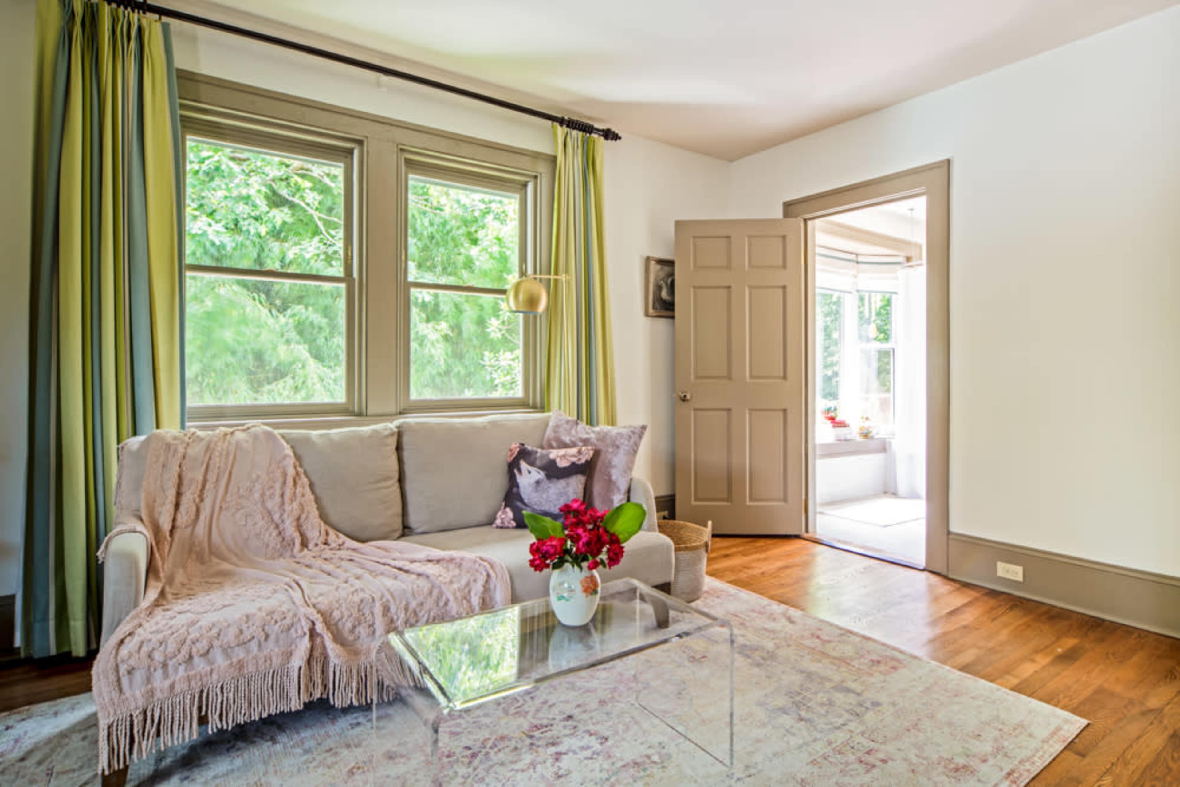 A light-colored living room features a sofa draped with a throw, a glass coffee table with flowers, and large windows providing natural light.
