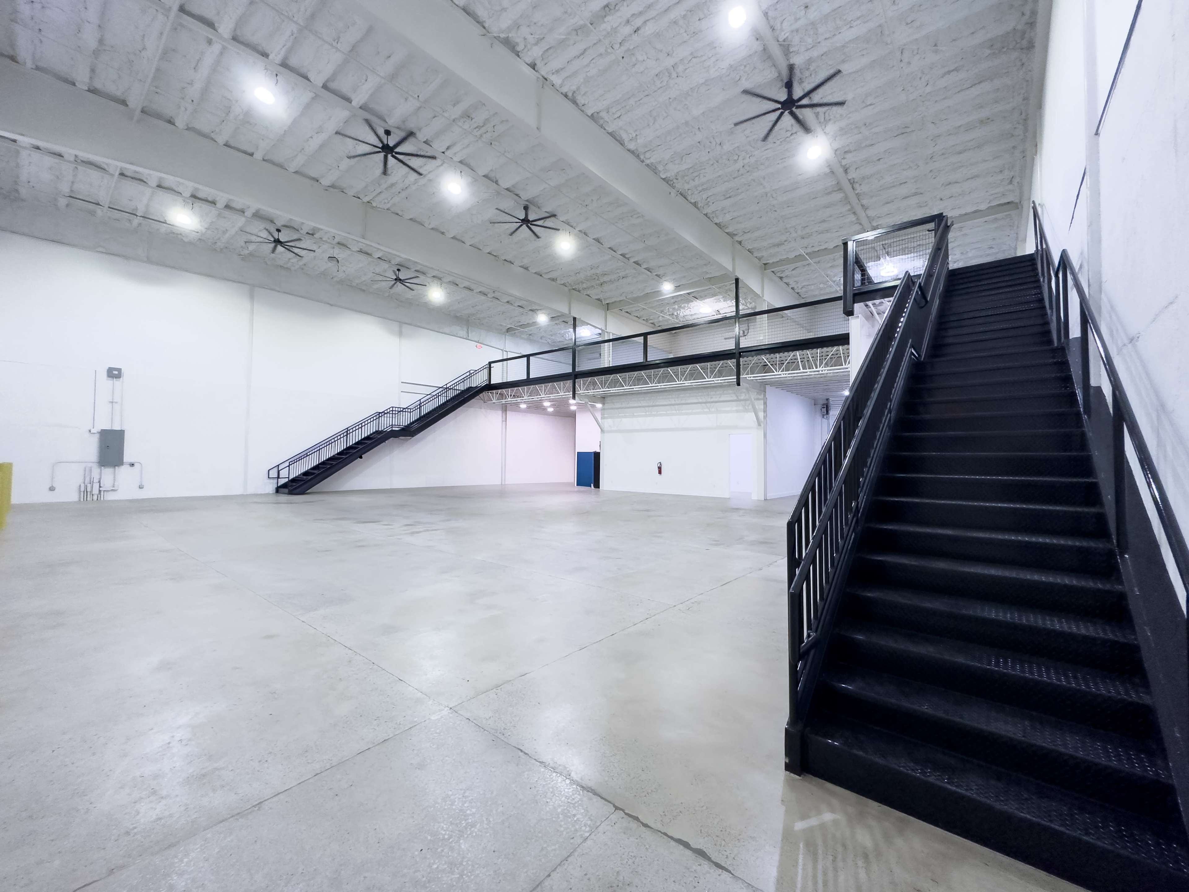The image shows a large, empty warehouse space with high ceilings, a staircase on the right leading to a mezzanine, and industrial ceiling fans.