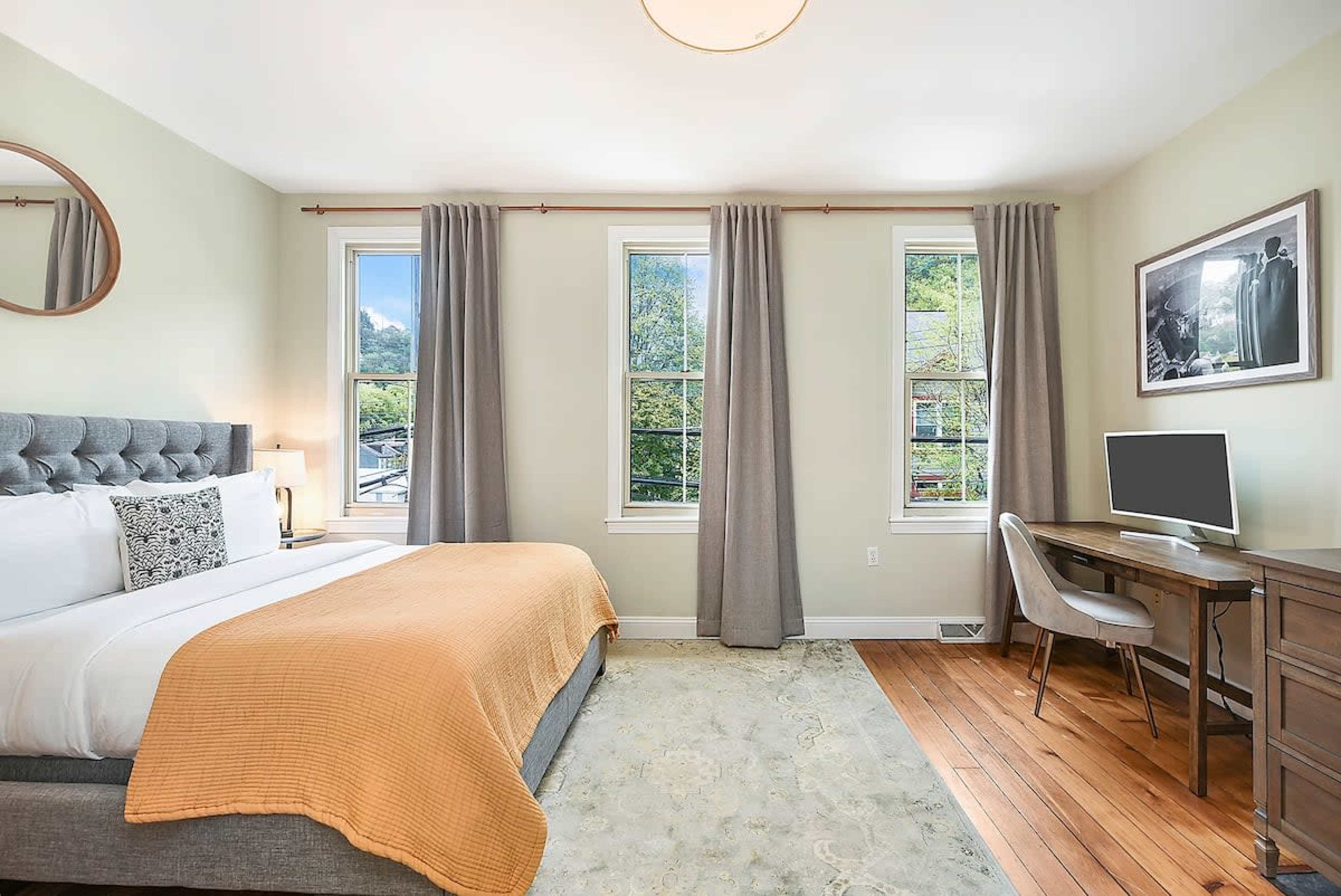 The bedroom features a gray upholstered bed with an orange throw blanket, a wooden desk with a chair, and large windows allowing natural light to fill the space.