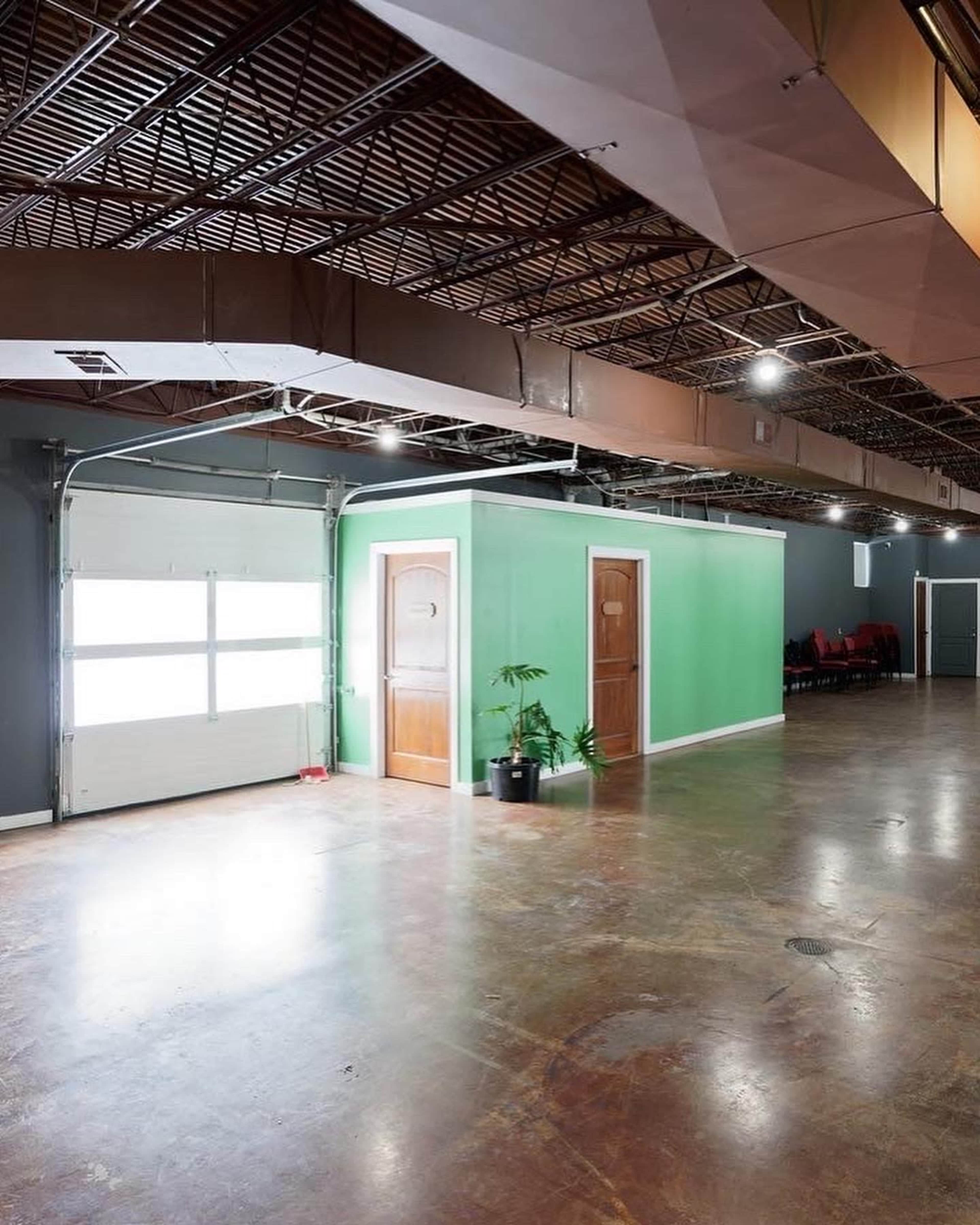 The image depicts a small green room with two wooden doors, situated in a large, empty industrial space with exposed ceiling and a concrete floor.