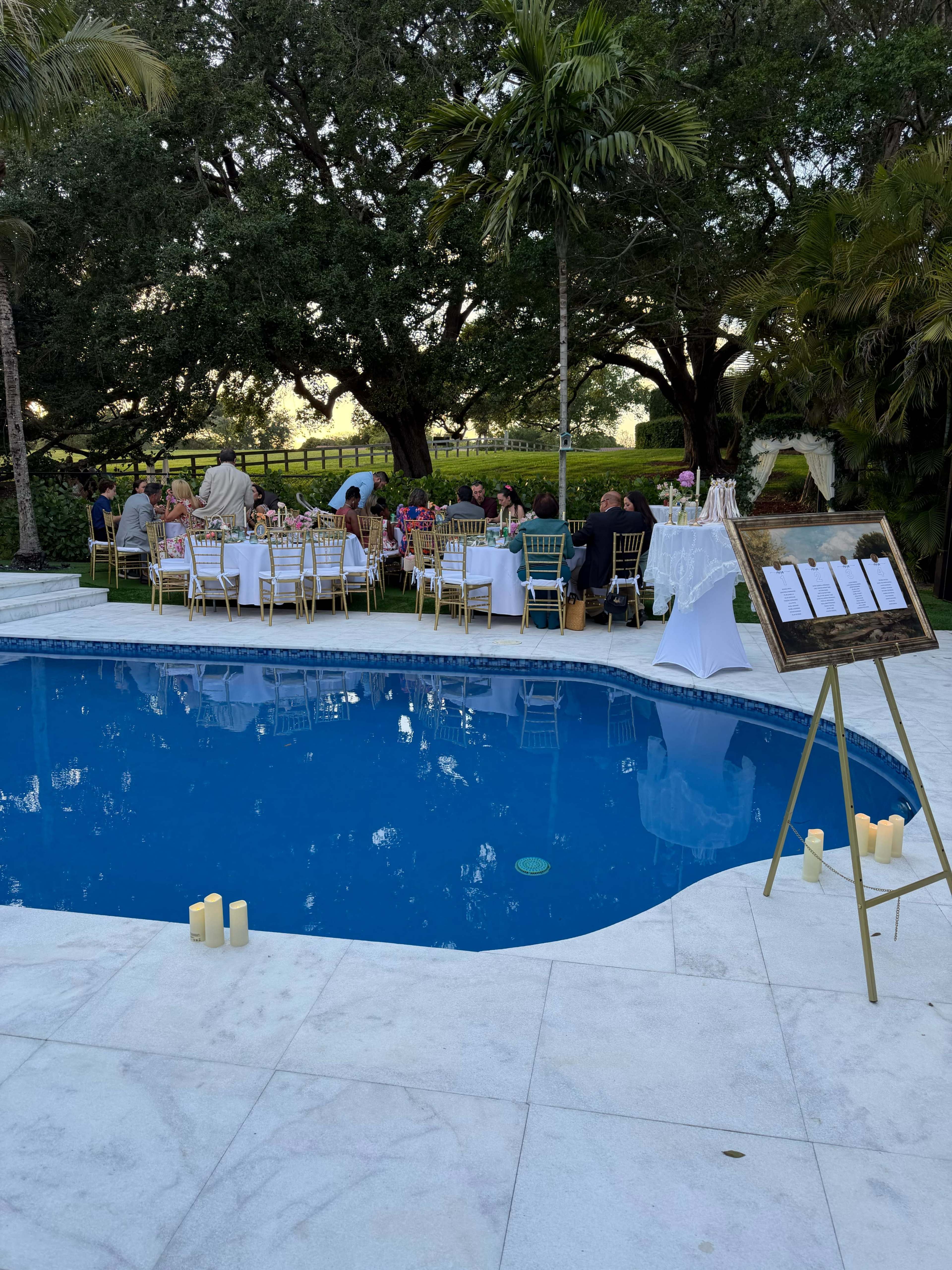 A dinner party is set up around a swimming pool, with tables arranged on the lawn under trees, and a display board featuring a list of names stands nearby.