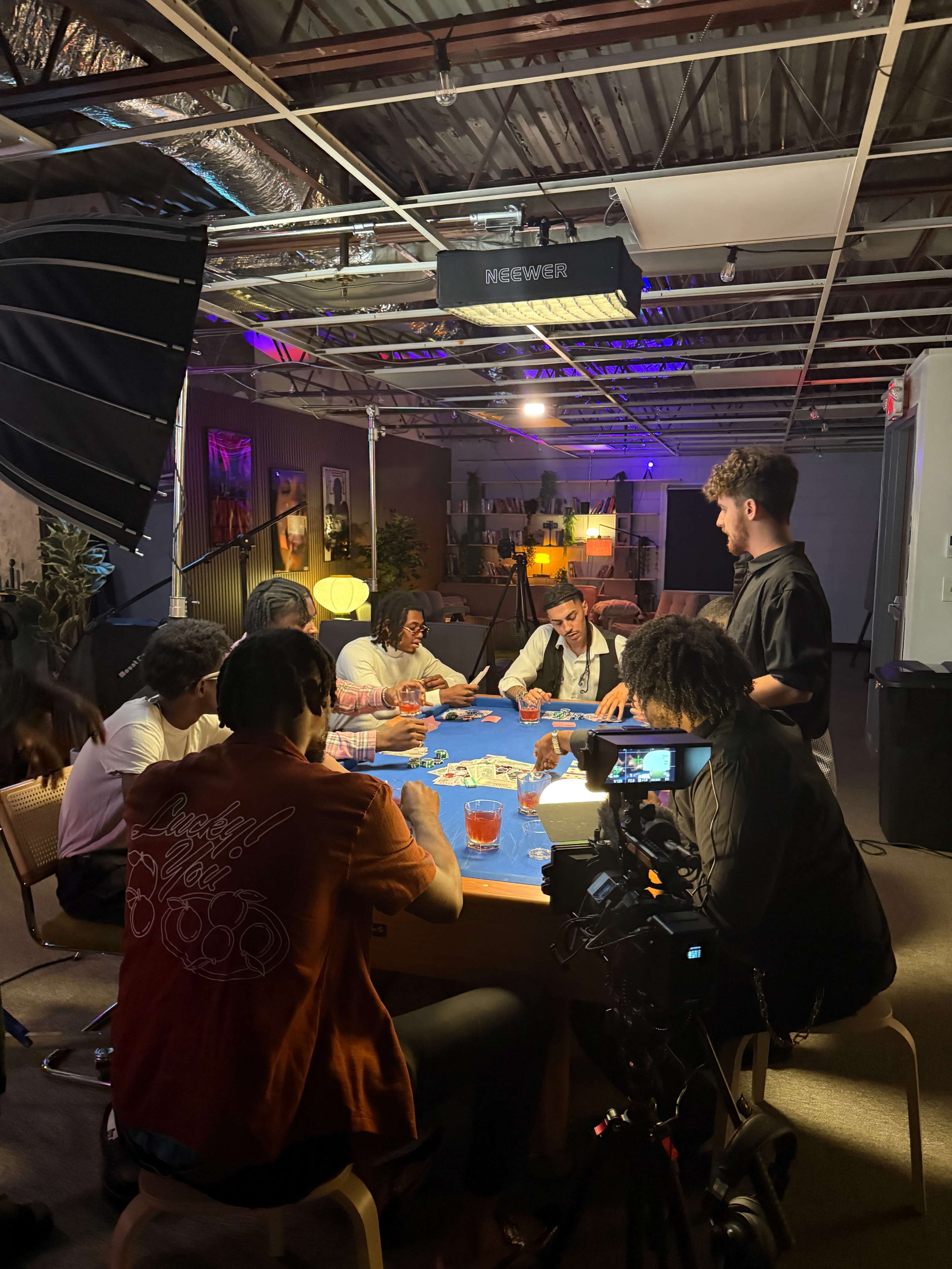 A group of people is seated around a table playing a card game, with cameras and lighting equipment set up in a well-lit indoor space.