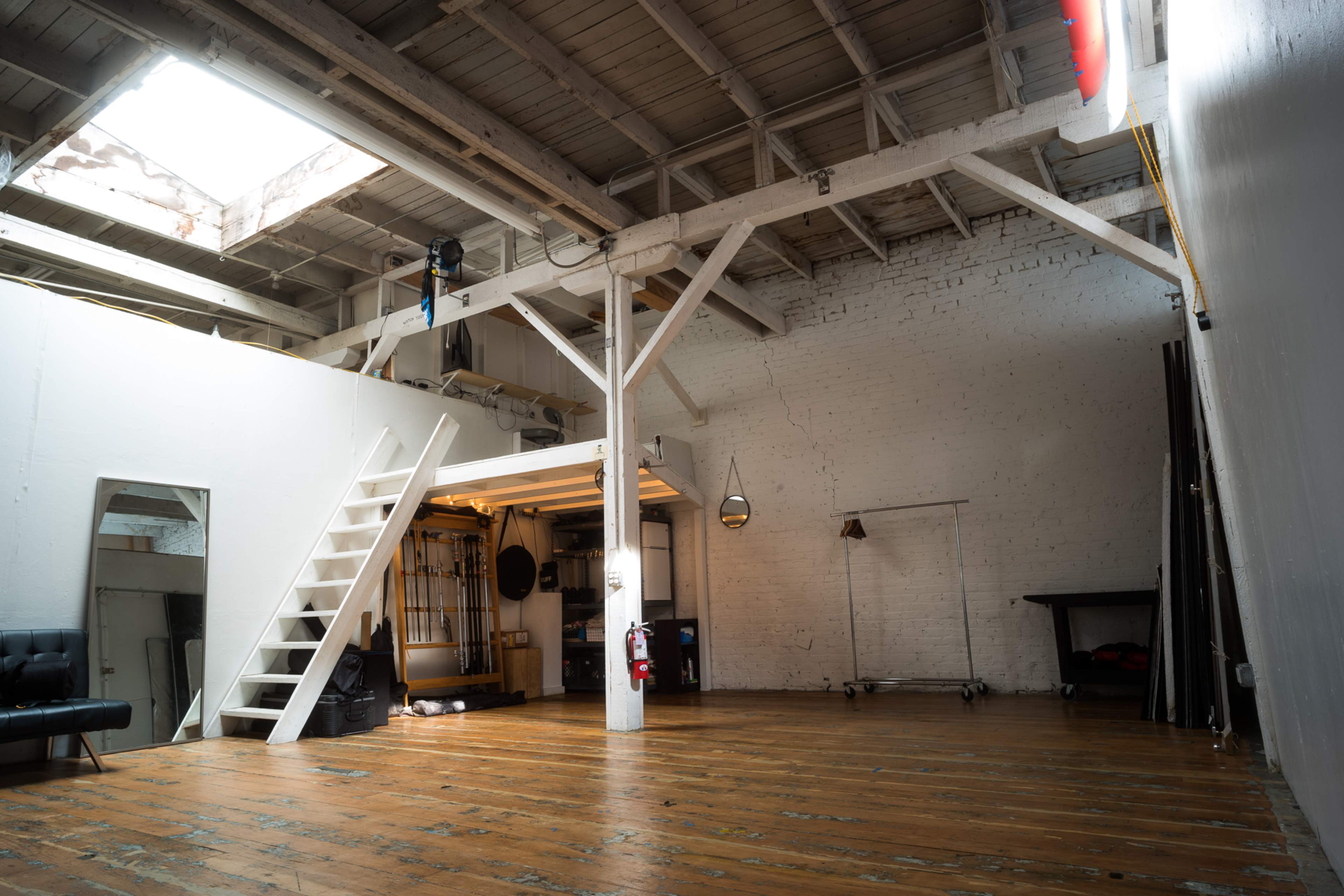 The image shows an open, spacious studio with wooden floors, exposed beams, and a ladder leading to a higher platform.