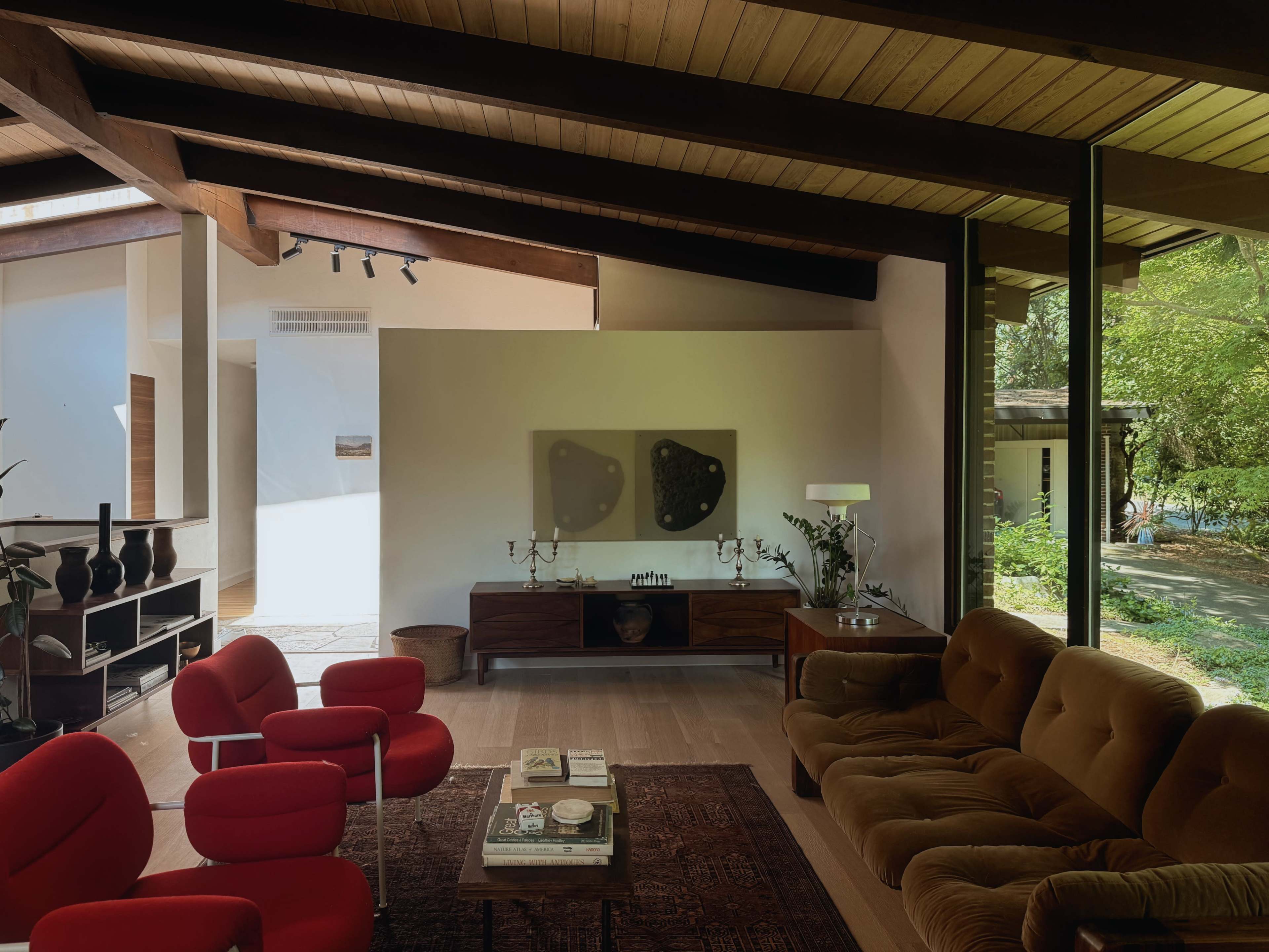 The image shows a modern living room featuring a brown sofa, red chairs, a wooden console, and large windows overlooking a green outdoor space.