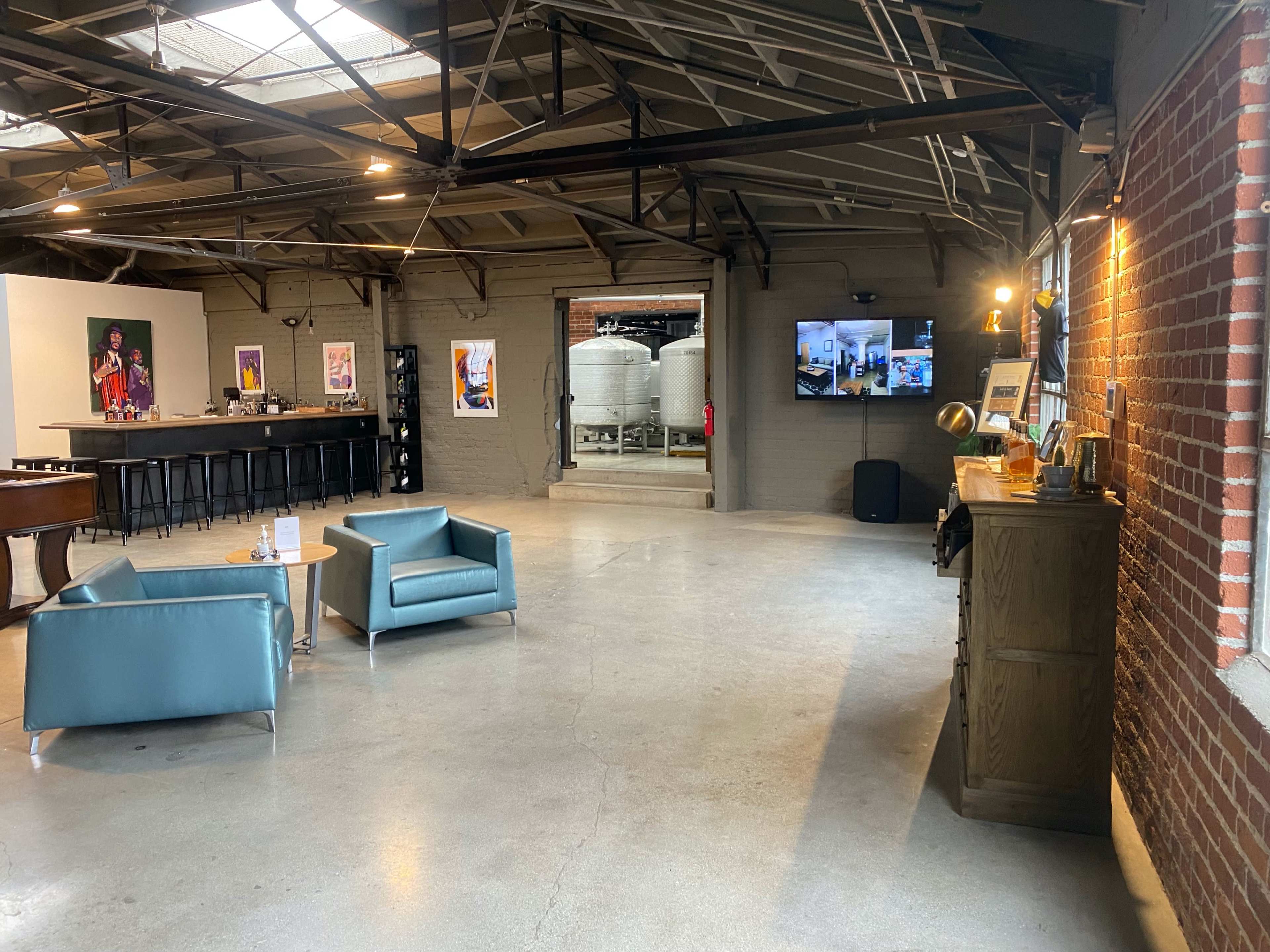 An industrial-style interior featuring a spacious area with two teal chairs, a wooden sideboard, and large fermentation tanks visible in the background.