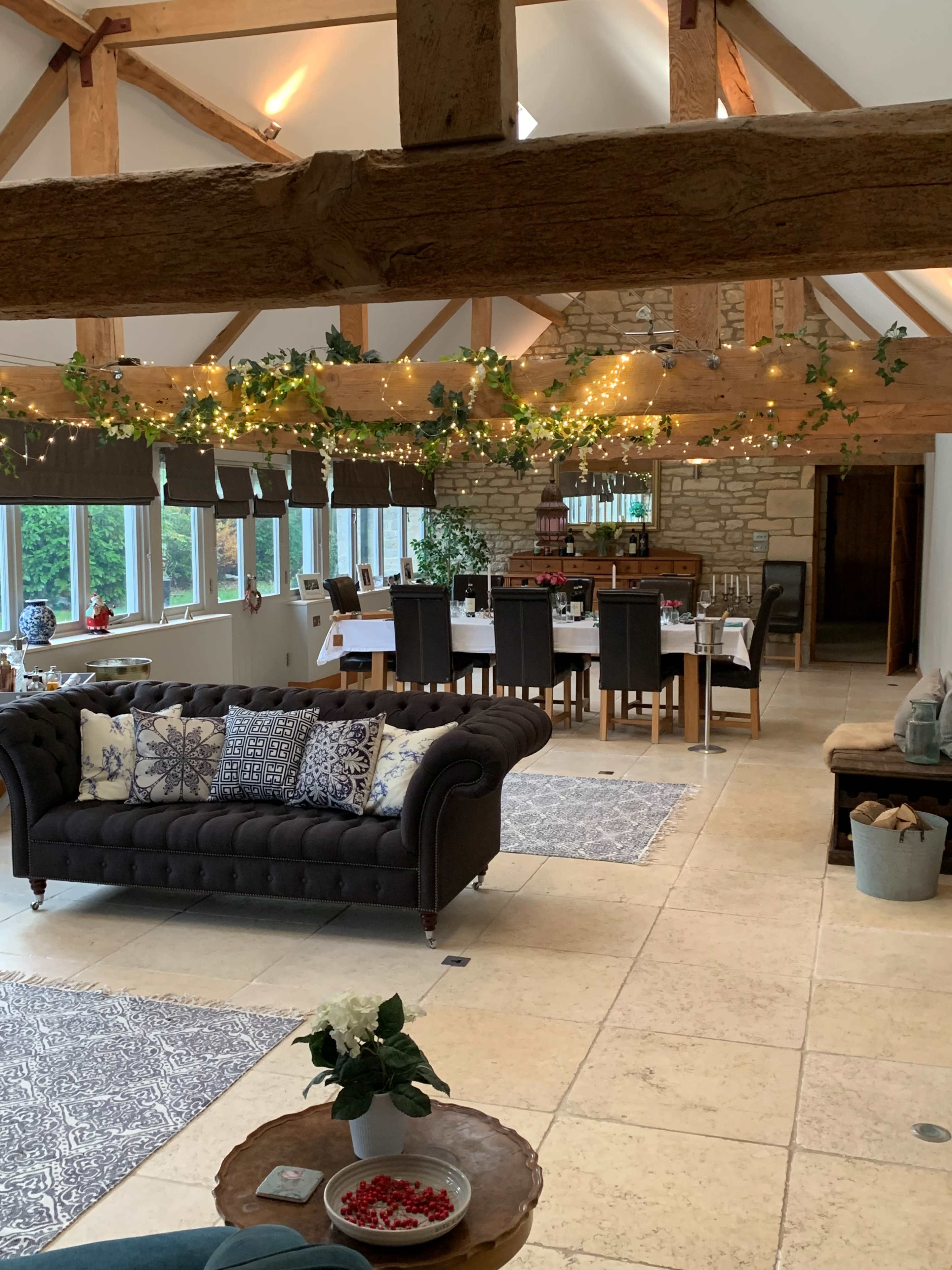 A spacious, open-plan room with a cozy seating area featuring a dark sofa, a dining table set for a meal, and wooden beams adorned with string lights.