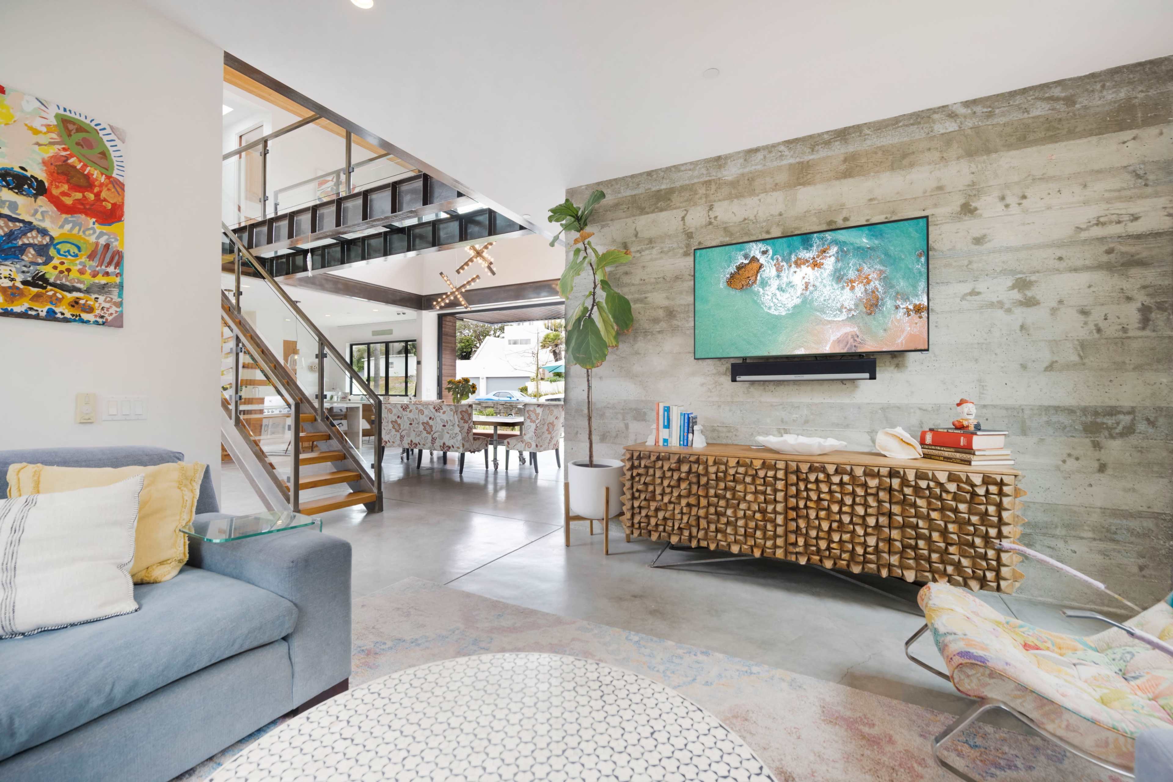 A modern living room with a concrete wall, a large television mounted on it, a patterned area rug, and a staircase leading to an upper level.