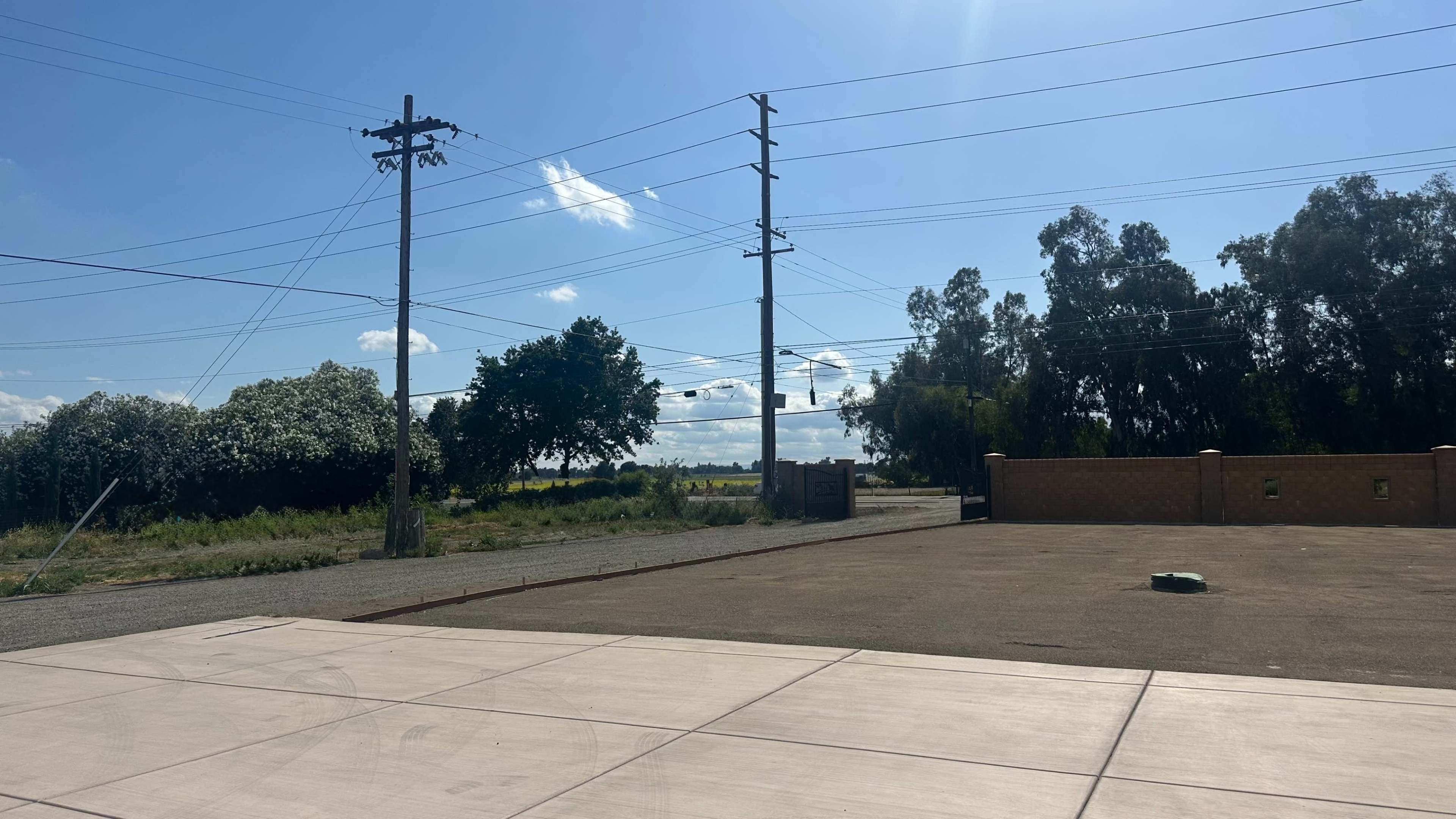 The image shows an empty lot with a concrete surface, utility poles, and trees in the background under a clear blue sky.