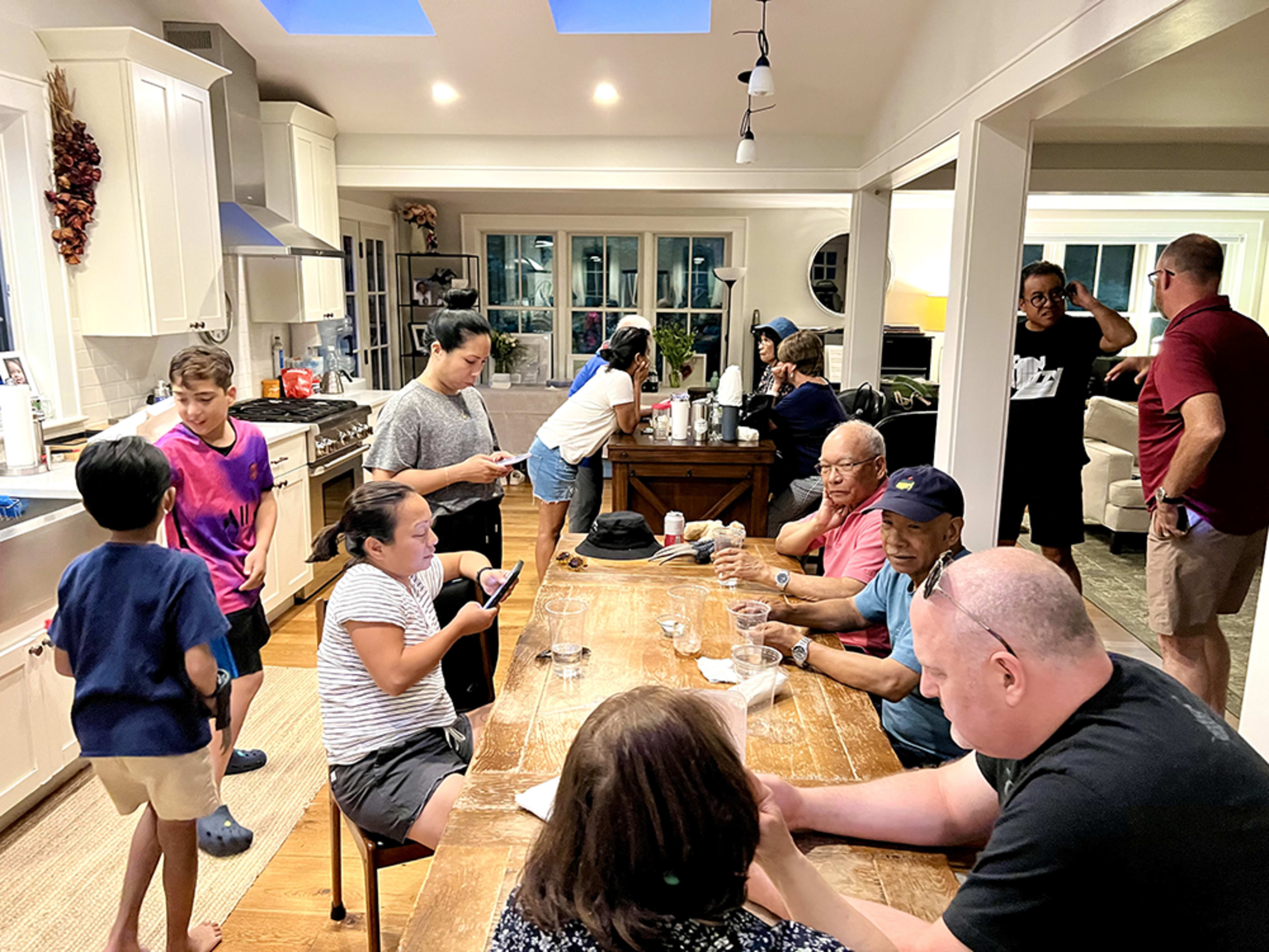 A group of people of various ages gathers around a large wooden table in a brightly lit kitchen, engaging in conversation and using smartphones.