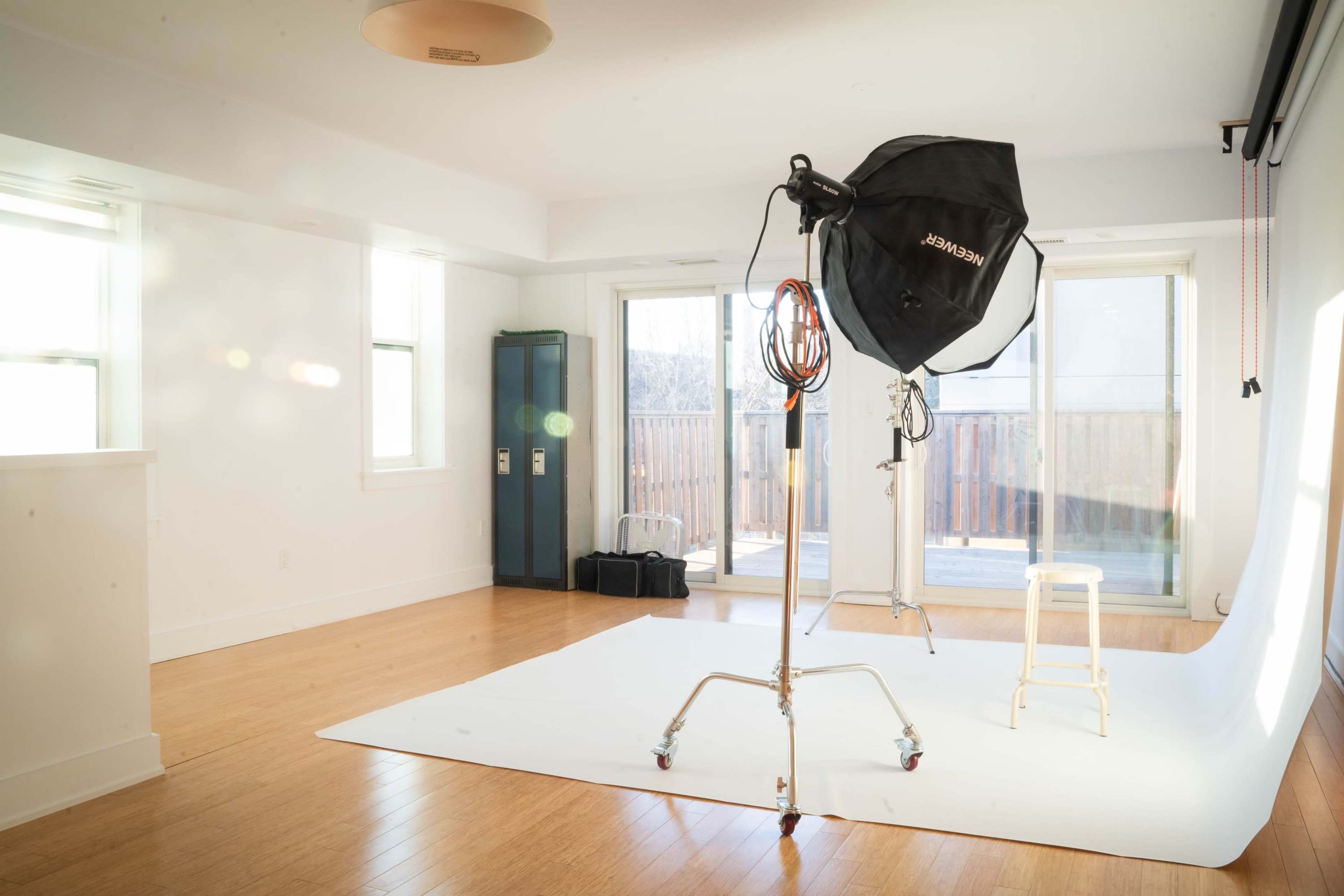 A photography studio features a white backdrop, a light stand with a softbox, and a stool, with large windows allowing natural light.