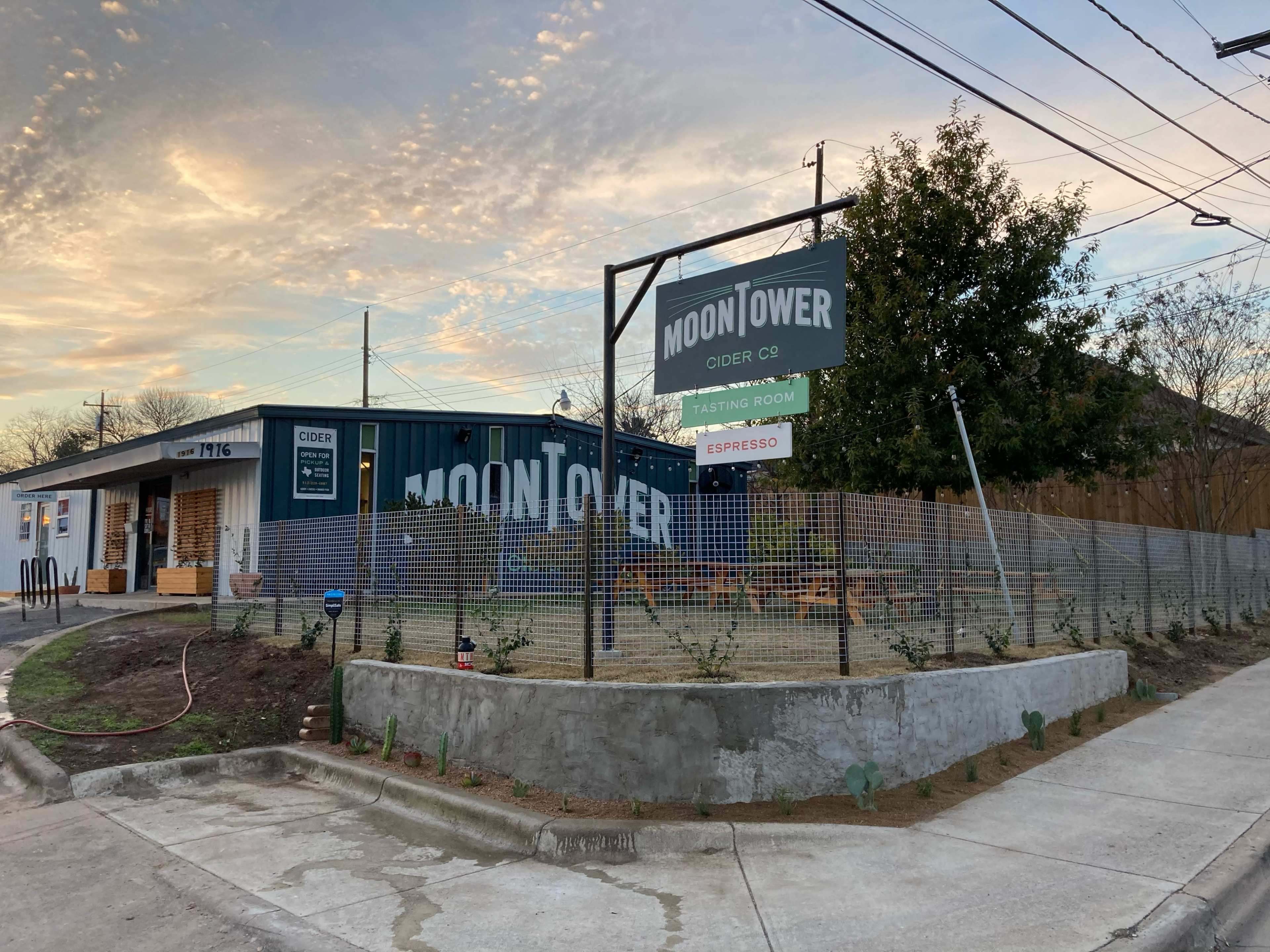 A building with a sign that reads "MOONTOWER CIDER CO." and "TASTING ROOM," surrounded by a landscaped area nearing completion at sunset.