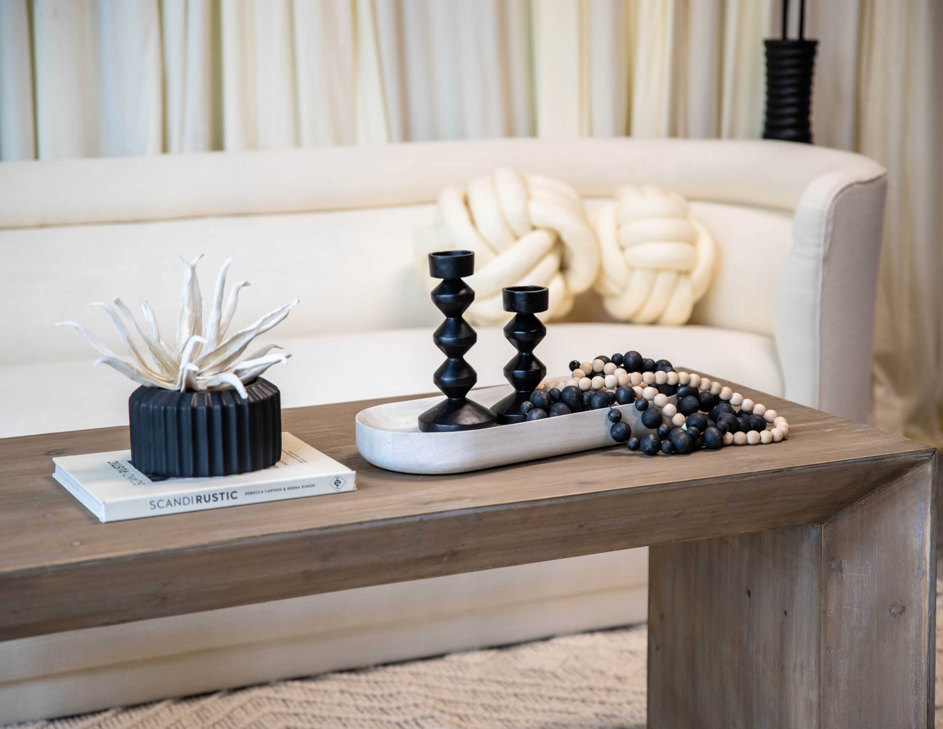 A wooden table is set with a decorative arrangement of candles, a textured bowl, and a beaded garland, in front of a cream-colored sofa.