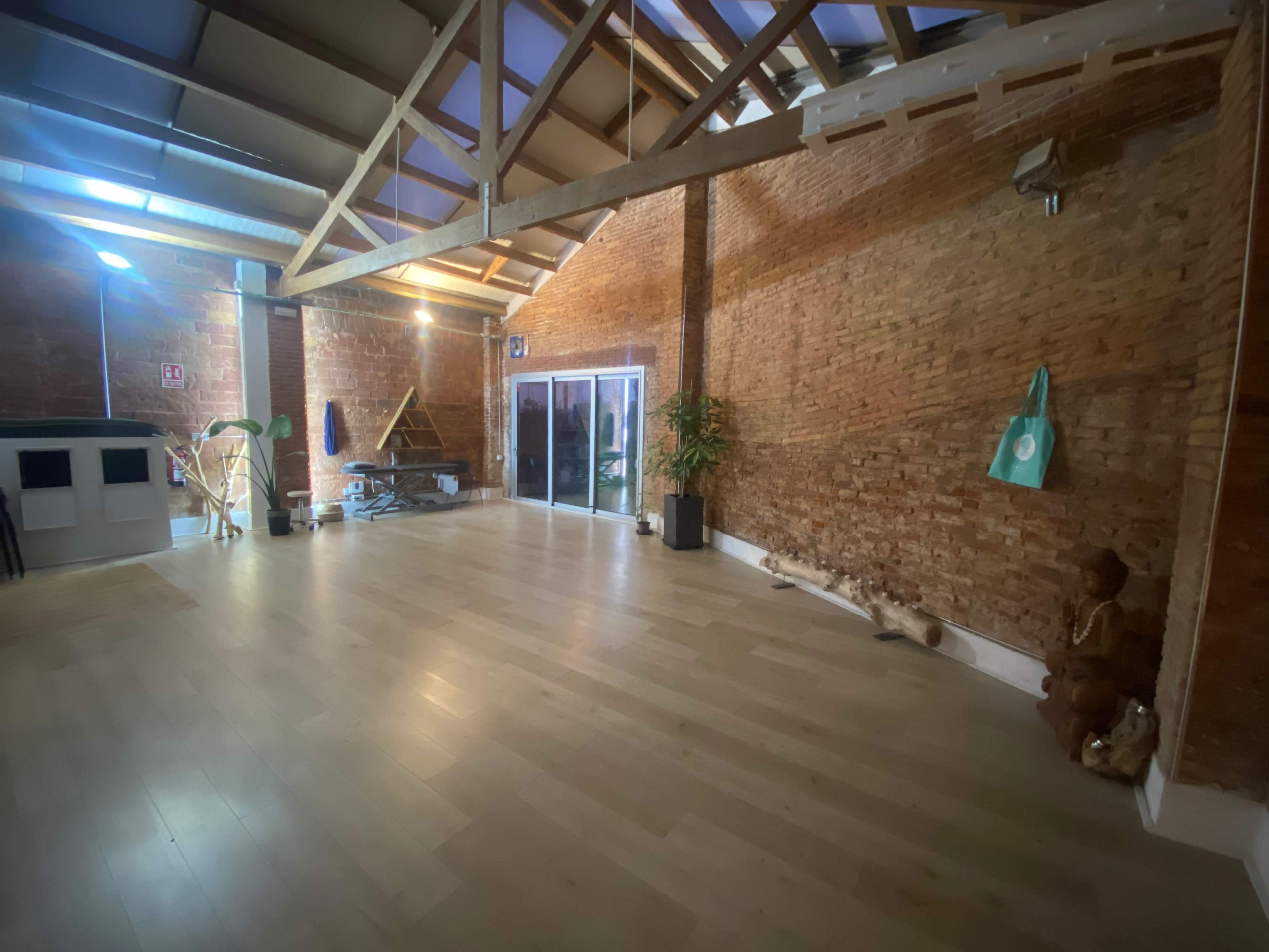 The image shows a spacious interior with exposed brick walls, wooden beams, and large glass doors leading outside, along with minimal furnishings.