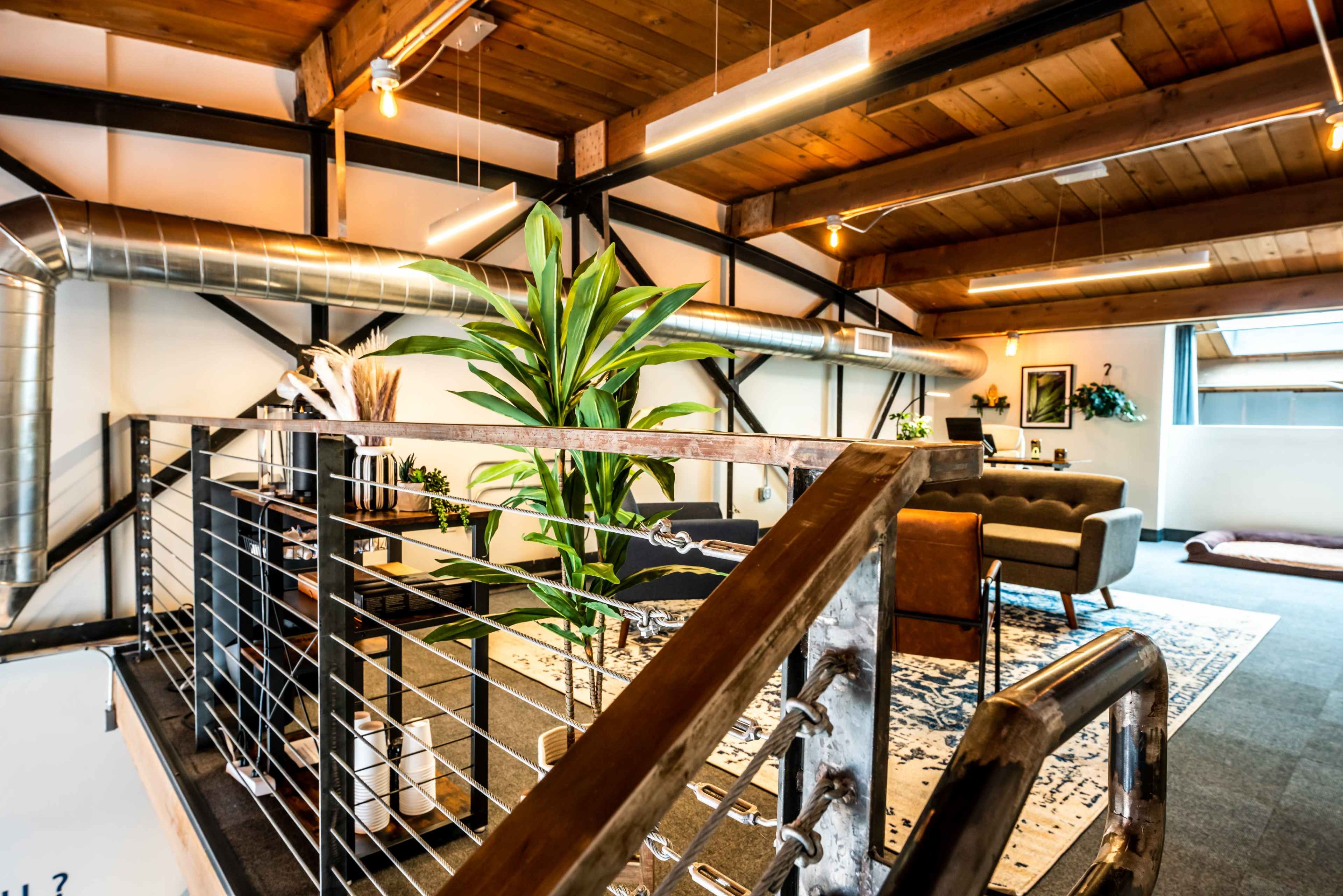 A stylish loft interior with wooden beams, metal accents, and a large potted plant near a railing overlooking a seating area.