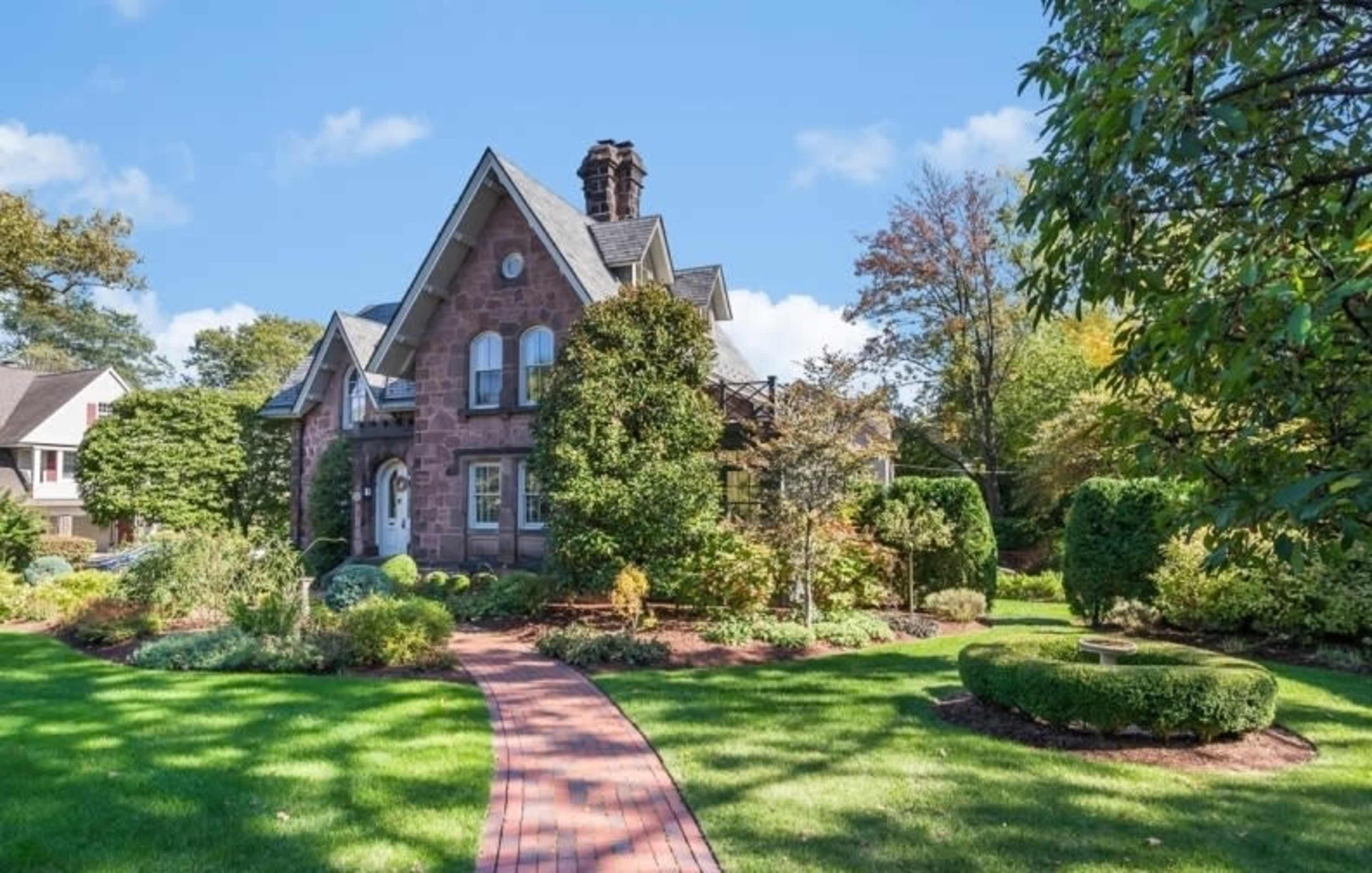 A two-story stone house with a steep roof and manicured gardens is surrounded by a brick pathway and lush greenery.