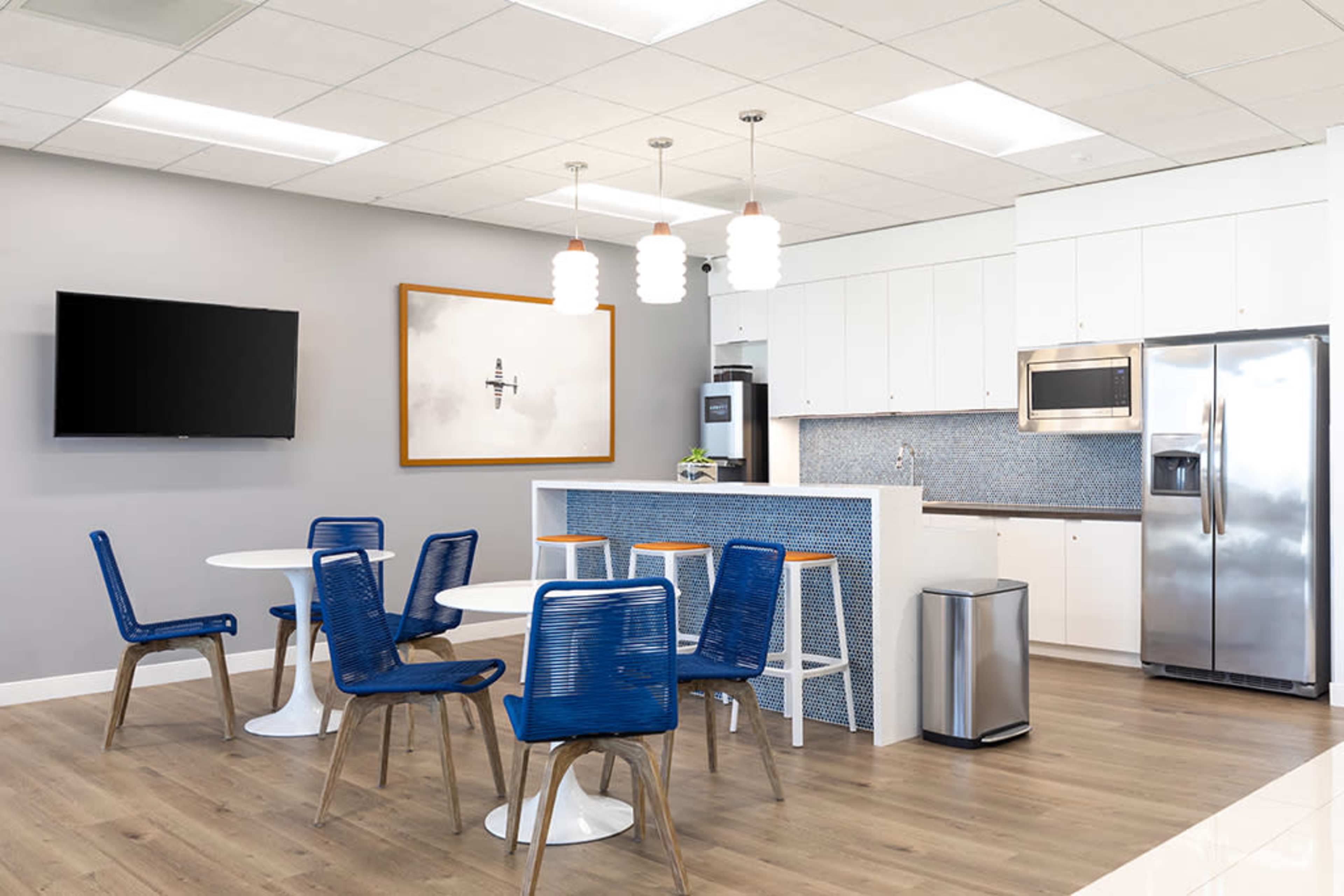 The image shows a modern kitchen area with a table and chairs, a refrigerator, a microwave, and a TV mounted on the wall.