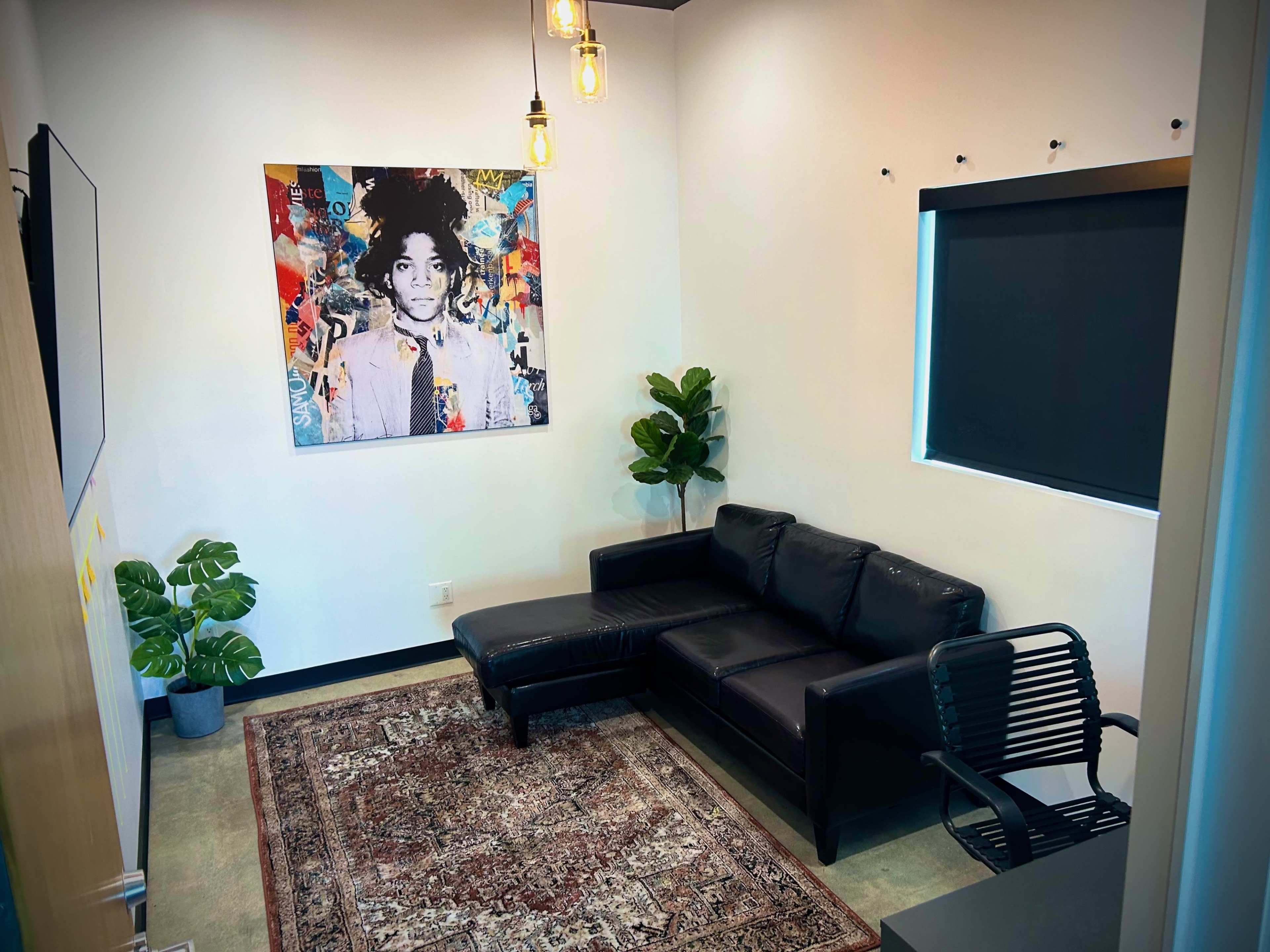 A small, modern room featuring a black leather sofa, a colorful artwork on the wall, a couple of indoor plants, and a chair near a window.