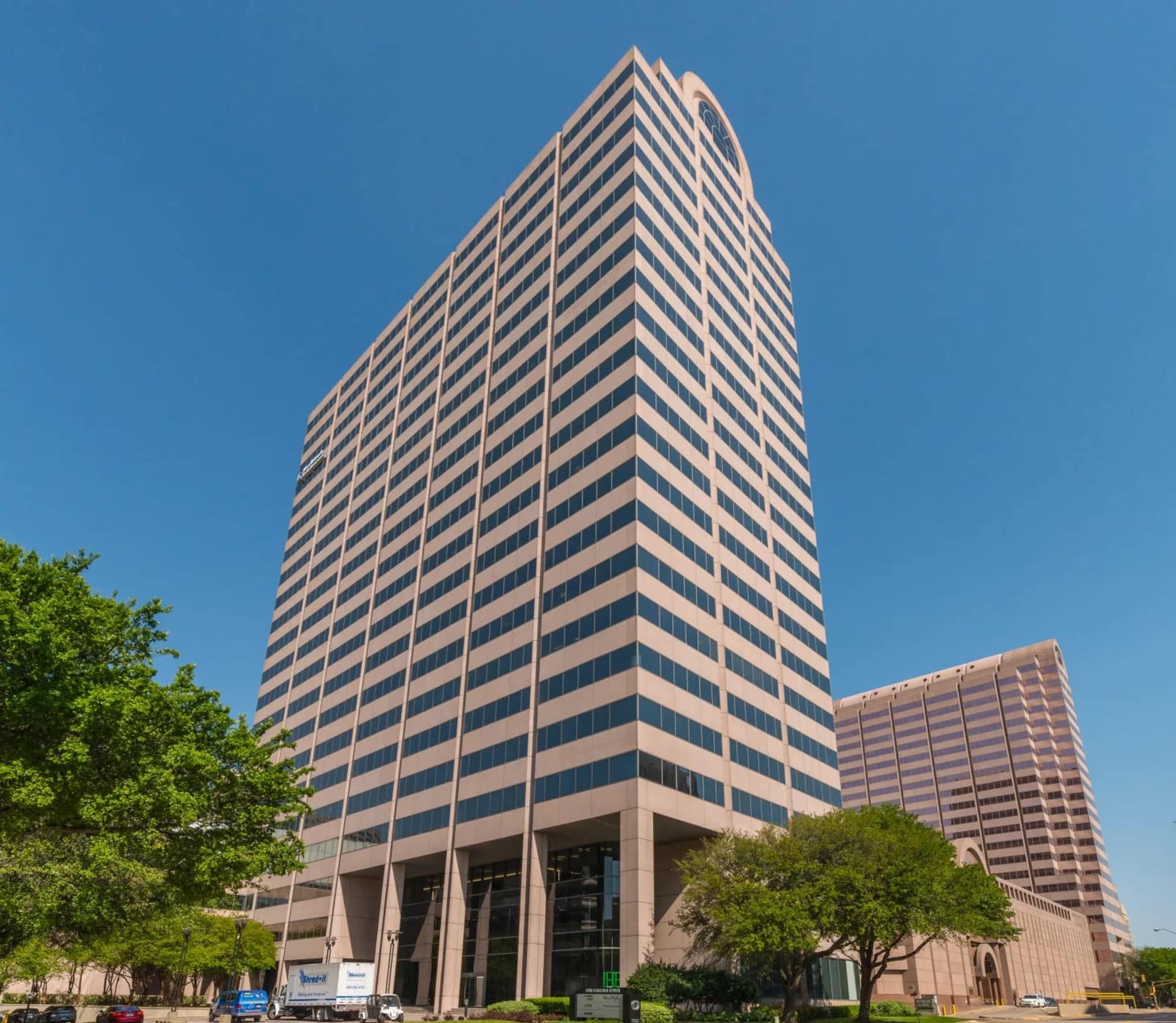 A tall office building with a modern design stands against a clear blue sky, flanked by trees and adjacent structures.