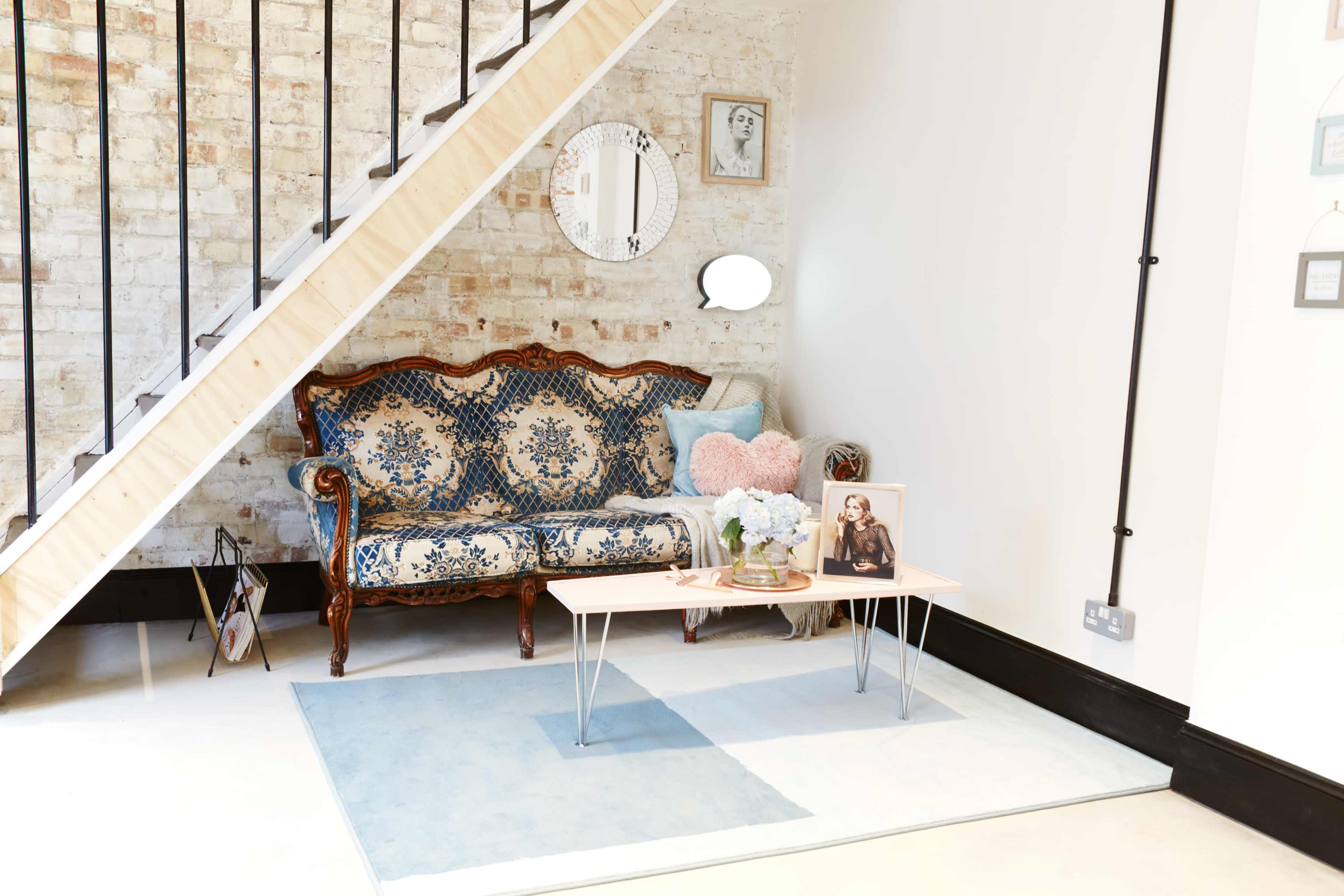 A vintage blue patterned sofa is positioned against a brick wall, accompanied by a coffee table adorned with a framed photo and a vase of flowers.